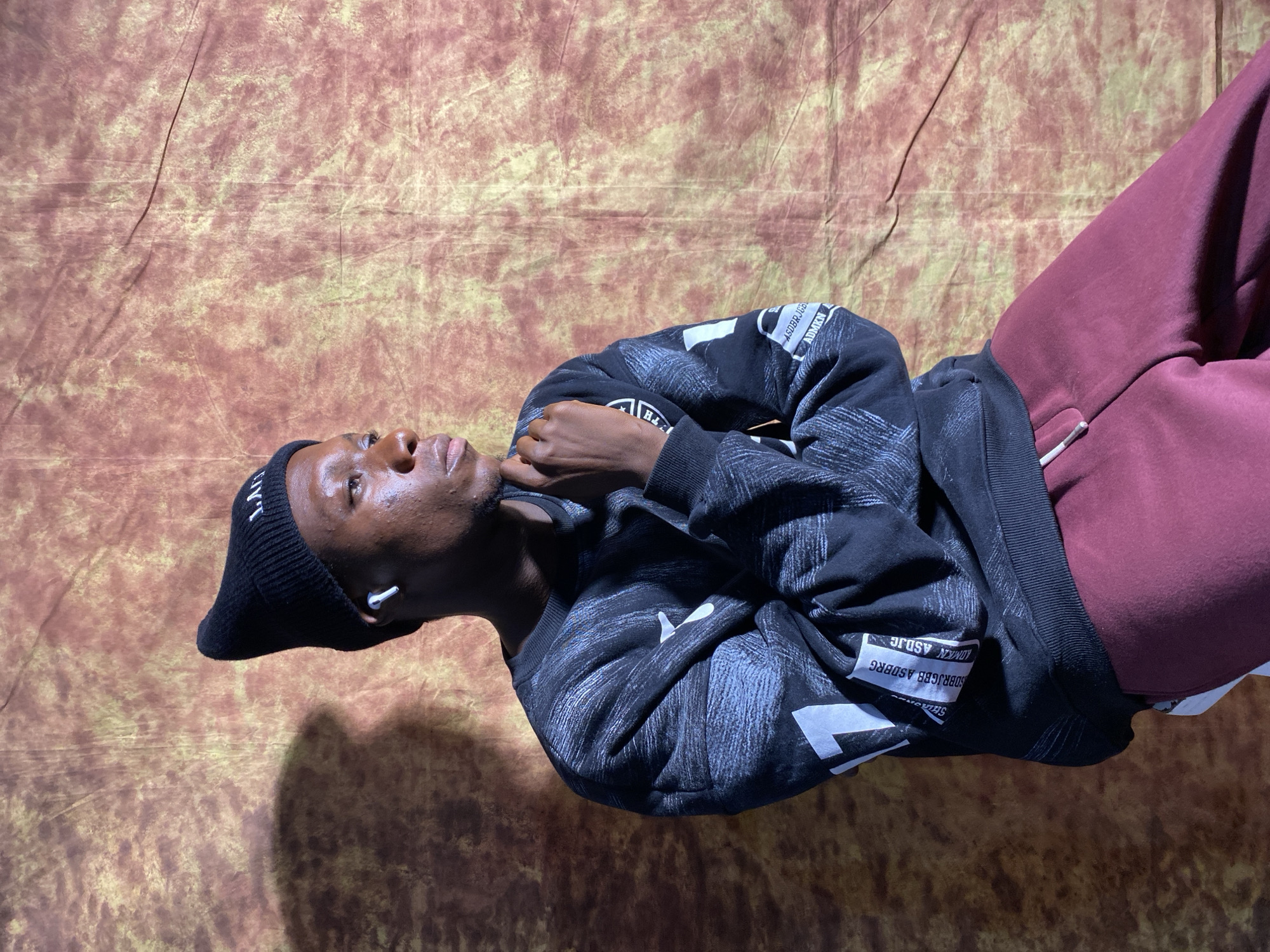In Barakin Akawo, Nigeria, a young Black male is seated against a textured, mottled backdrop that appears to be a fabric backdrop. He is wearing a black beanie with white lettering, a black sweatshirt with a distressed pattern and white graphics on the sleeves, and maroon sweatpants. He has white wireless earbuds in his ears and is looking to his right, with his chin resting on his hand. A shadow is cast on the backdrop to his left.