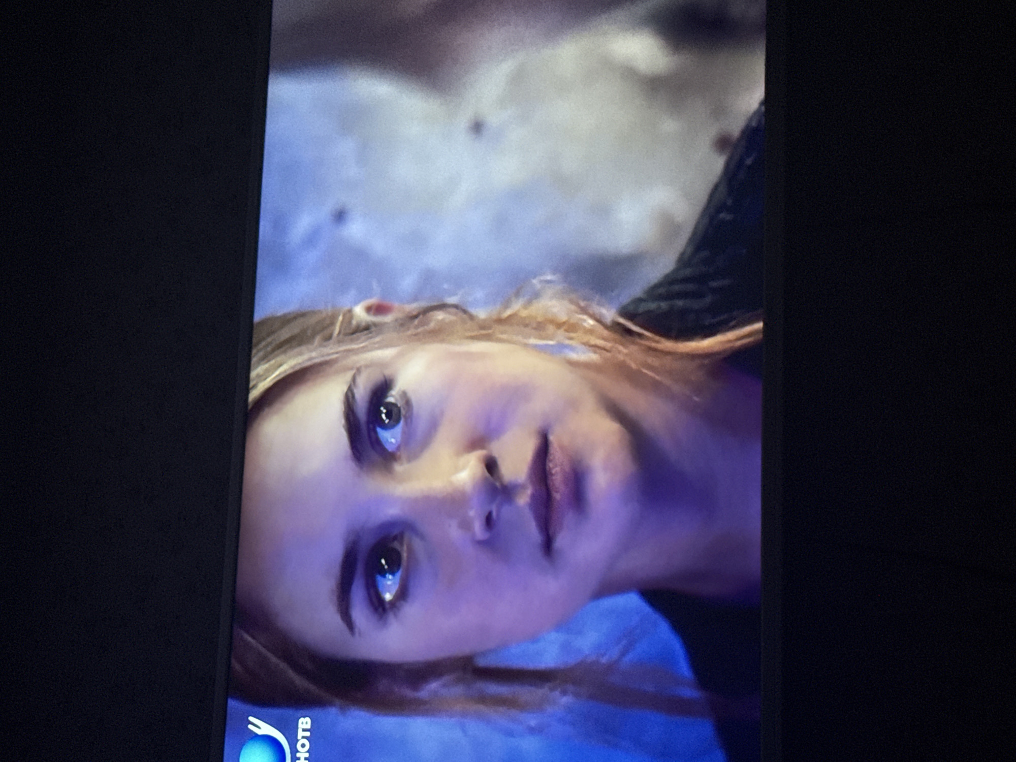 The image is a close-up shot of a fair-skinned woman with light brown hair. Her face is angled towards the camera, with her eyes looking directly at the viewer. The lighting is predominantly blue and purple, casting shadows across her face. She has noticeable eyebrows and dark mascara. Her lips are slightly parted. The bottom left corner of the image features a logo that reads "HOTB." The background is blurred and out of focus, but appears to be a mix of blue and gray tones. The photo seems to be a screenshot from a television or monitor, evident from the black edges on either side. The image doesn't provide any information about the city of Mytishchi, Russia, or any specific location.