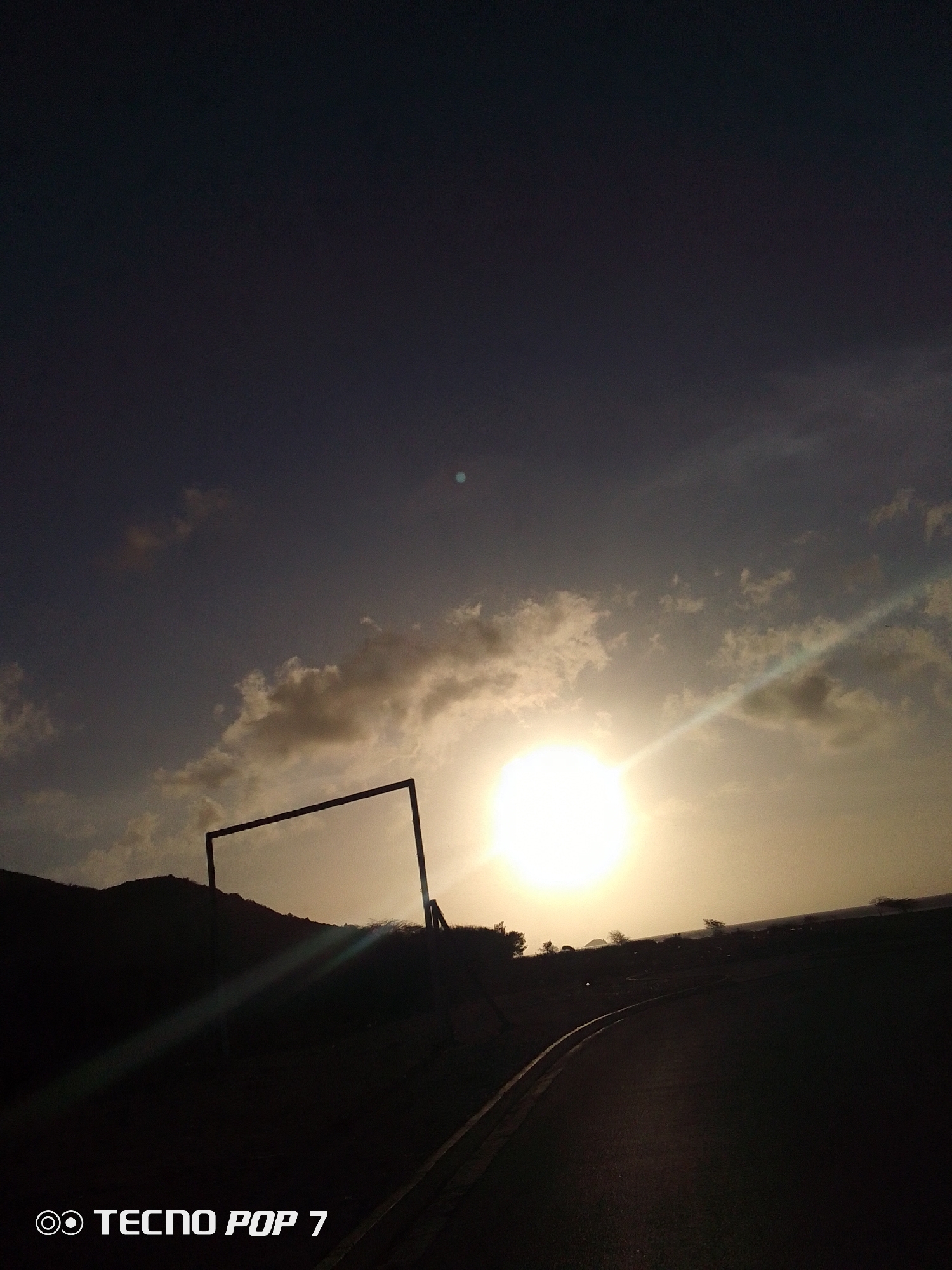 The image shows a sunset scene with a dark sky at the top transitioning to a lighter, cloudier sky near the horizon. The sun is a bright, central point, with sun rays emanating from it. In the foreground, there is a dark road curving to the right and a dark rectangular frame standing near the horizon. The landscape is silhouetted, with a dark hill and some trees visible. The text "TECNO POP 7" is displayed at the bottom of the image. The location is Pedro González, Venezuela.