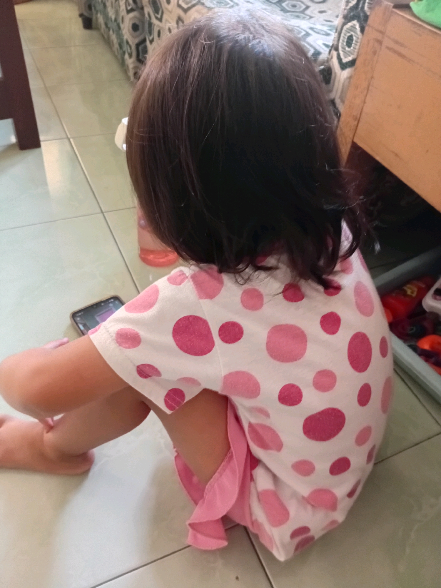 A young child with dark hair is sitting on a tiled floor, facing away from the camera. The child is wearing a white shirt with pink polka dots and a pink skirt. A smartphone is visible in the child's left hand. The background shows a portion of a patterned couch and a wooden piece of furniture. The scene appears to be indoors, likely in a residential setting within Gebang, Indonesia.