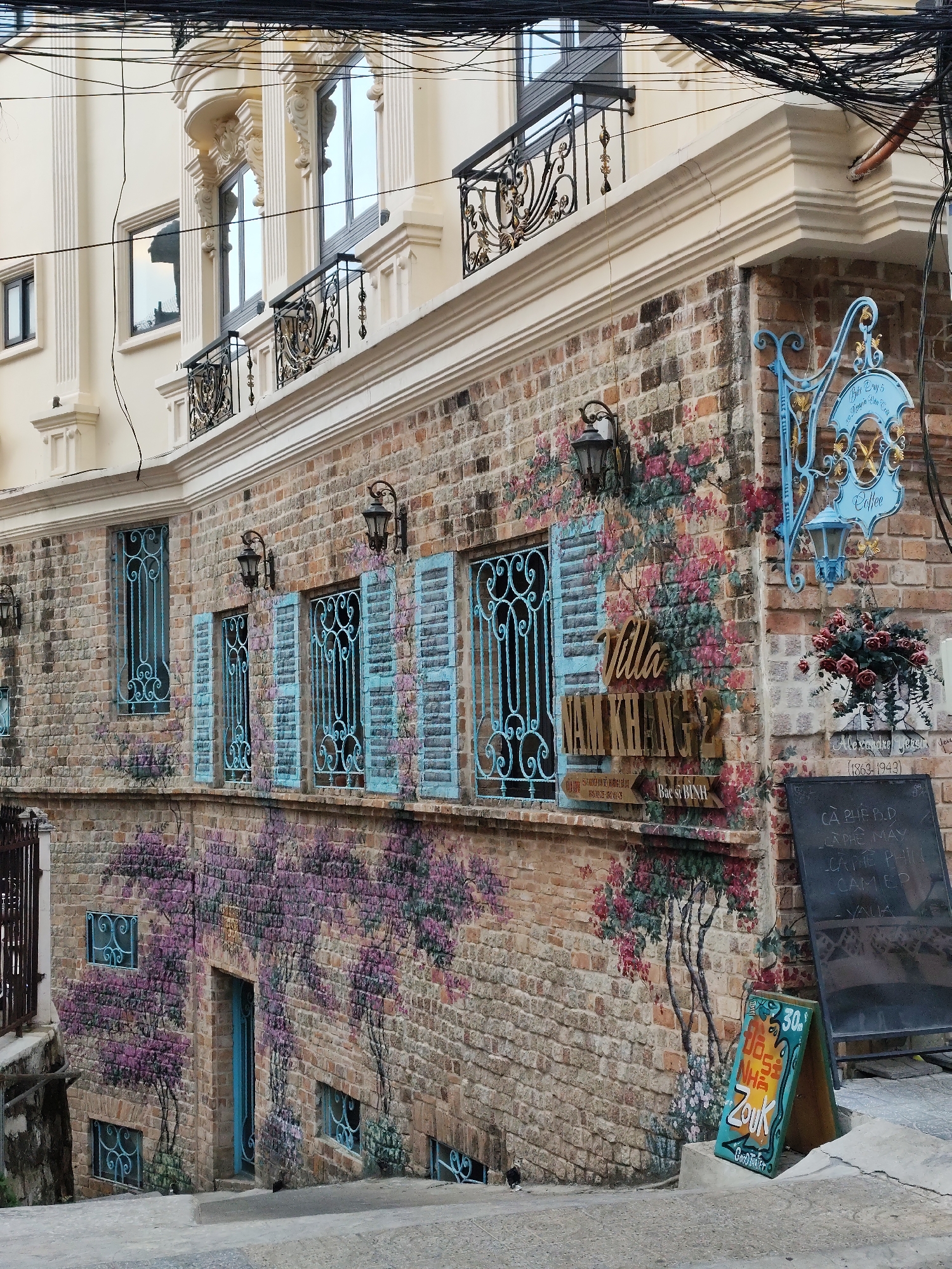 This image shows the Villa Nam Khang cafe in Da Lat, Vietnam. The building's exterior is brick, adorned with floral paintings, light blue wrought-iron window coverings, and hanging flower baskets. Signage includes "Villa Nam Khang" and a board listing coffee offerings in Vietnamese. Additional signage in blue and red advertises the cafe. An upper floor with ornate balconies is visible. Power lines are visible overhead.