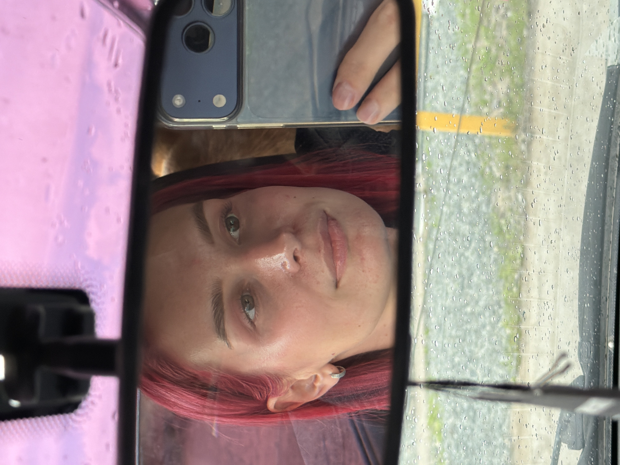 An upside-down close-up shot captures the reflection of a person in a car's rearview mirror, facing the viewer. The individual, likely a young female, has prominent red or magenta-colored hair that frames their face and partially covers their shoulder. Their eyes, a light shade of brown or green, are open and look directly towards the mirror's perspective. The person's expression is neutral, with slightly parted lips. A small, silver hoop earring is visible in their left ear.
A hand, presumably the person's, is visible holding a smartphone with a blue protective casing, which features a dual-camera setup and a flash. The phone is positioned above the person's head, partially obscuring the top of their forehead and hair in the reflection.
The background visible through the wet car windshield, outside the mirror's frame and to the right, shows numerous rain droplets, indicating recent or ongoing precipitation. Beyond the water-streaked glass, a blurry outdoor scene reveals green vegetation and a horizontal yellow line, possibly a road marking or curb, suggesting an outdoor parking lot or roadside setting. The left side of the reflection, outside the mirror, shows a pinkish or purplish hue, possibly from tinted glass or an object inside the car. The overall setting appears to be inside a vehicle during daytime in Kamyennoye, Russia, with overcast or rainy weather conditions. No discernible text is visible in the image.