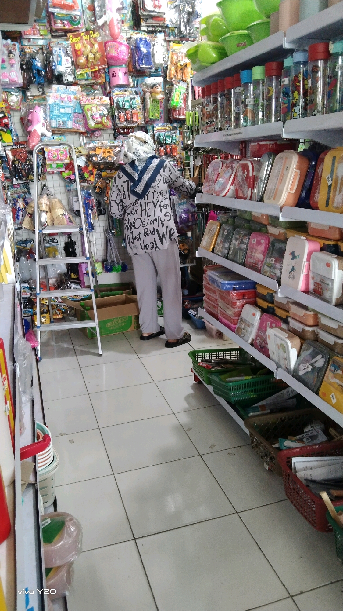 In a retail store located in Karawang, Indonesia, a person wearing a white hijab and a patterned shirt is standing and facing away from the camera. The person is also wearing light gray pants and dark slippers. The store is filled with shelves stacked high with various items, including what appear to be food containers, water bottles, and toys. A silver ladder stands to the left of the person. The floor is tiled in a light color.