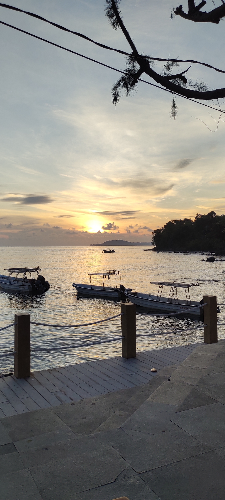 The image depicts a serene sunset scene at a pier in Sabang, Indonesia. The sky is painted with warm hues of orange and yellow as the sun dips below the horizon, casting a soft glow on the water. Several boats are moored at the pier, their silhouettes dark against the vibrant sky. The water is calm, reflecting the colors of the sunset. In the foreground, the wooden planks of the pier are visible, leading the viewer's eye towards the water. The overall atmosphere is peaceful and tranquil, a perfect representation of a tropical evening.