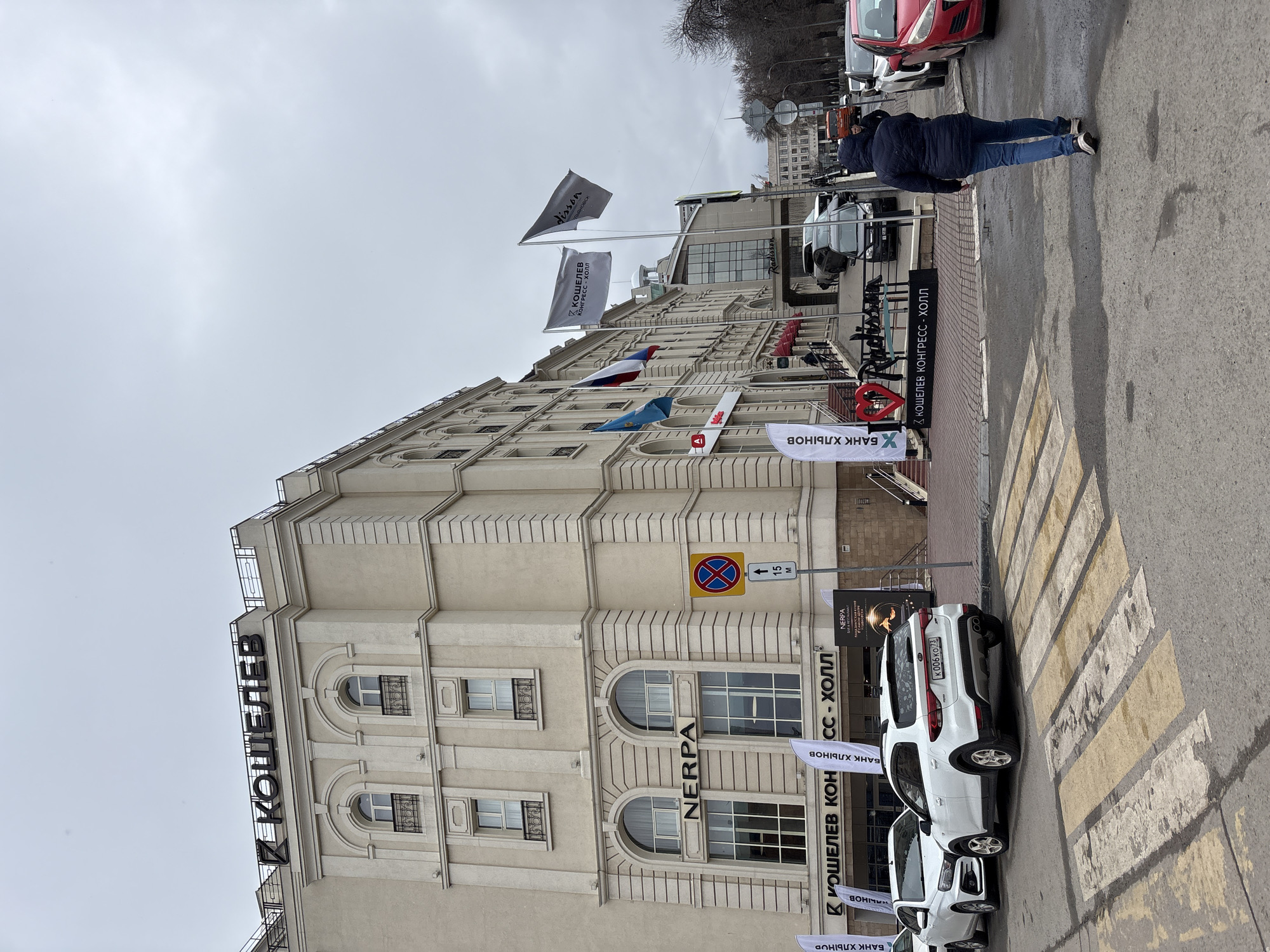 Here is a description of the image:
The image shows the "Кошелев Конгресс-Холл" hotel in Ulyanovsk, Russia. The building is a light-colored, multi-story structure with arched windows and ornate architectural details. A vertical sign on the building spells out "КОШЕЛЕВ" (Koshelev).
Several flags are displayed near the entrance, including the Russian flag, the flag of the Ulyanovsk region, and flags related to the hotel or associated businesses, one of them including "disson Ulyanovsk." A sign indicates a no-stopping zone with a distance of 15 meters.
Two white cars are parked near the building, on a pedestrian crossing. A person in a dark jacket and jeans is seen walking away from the camera on the sidewalk. The sky is overcast, and the overall scene appears to be an urban street setting. Street-level signage is visible promoting "Банк Хлынов" (Bank Khlynov) and "Nerpa."