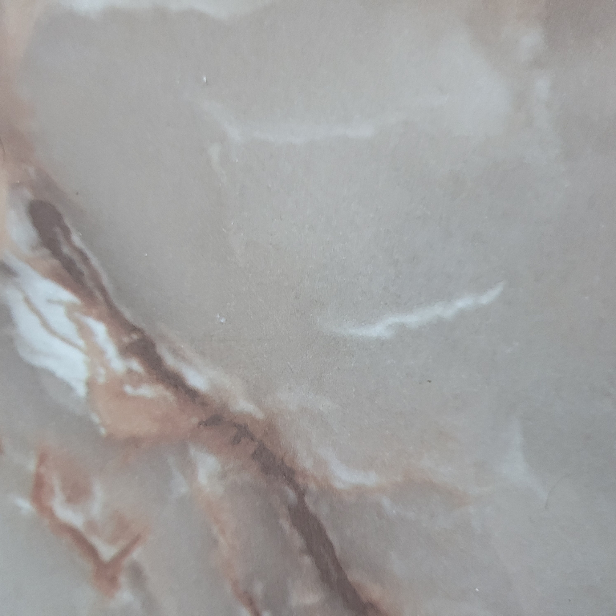 The image displays a close-up, abstract view of a surface, likely a natural stone such as marble or a marble-effect synthetic material. The dominant colors are light beige and pale grey, serving as the base tone. Interspersed throughout this lighter background are intricate, irregular patterns of darker reddish-brown and contrasting bright white veining. The reddish-brown elements appear as flowing, organic streaks and broader, diffuse patches, suggesting natural mineral deposits. The white veins are typically thinner and more sharply defined, cutting across the surface in wavy or linear patterns. The surface appears smooth, possibly polished, and exhibits a very fine, subtle texture. No people, objects, or specific events are depicted. The lighting is even, illuminating the surface without casting discernible shadows. There are no visible text elements or specific location cues within the image itself, though it is contextualized as being from Yerevan, Armenia.
