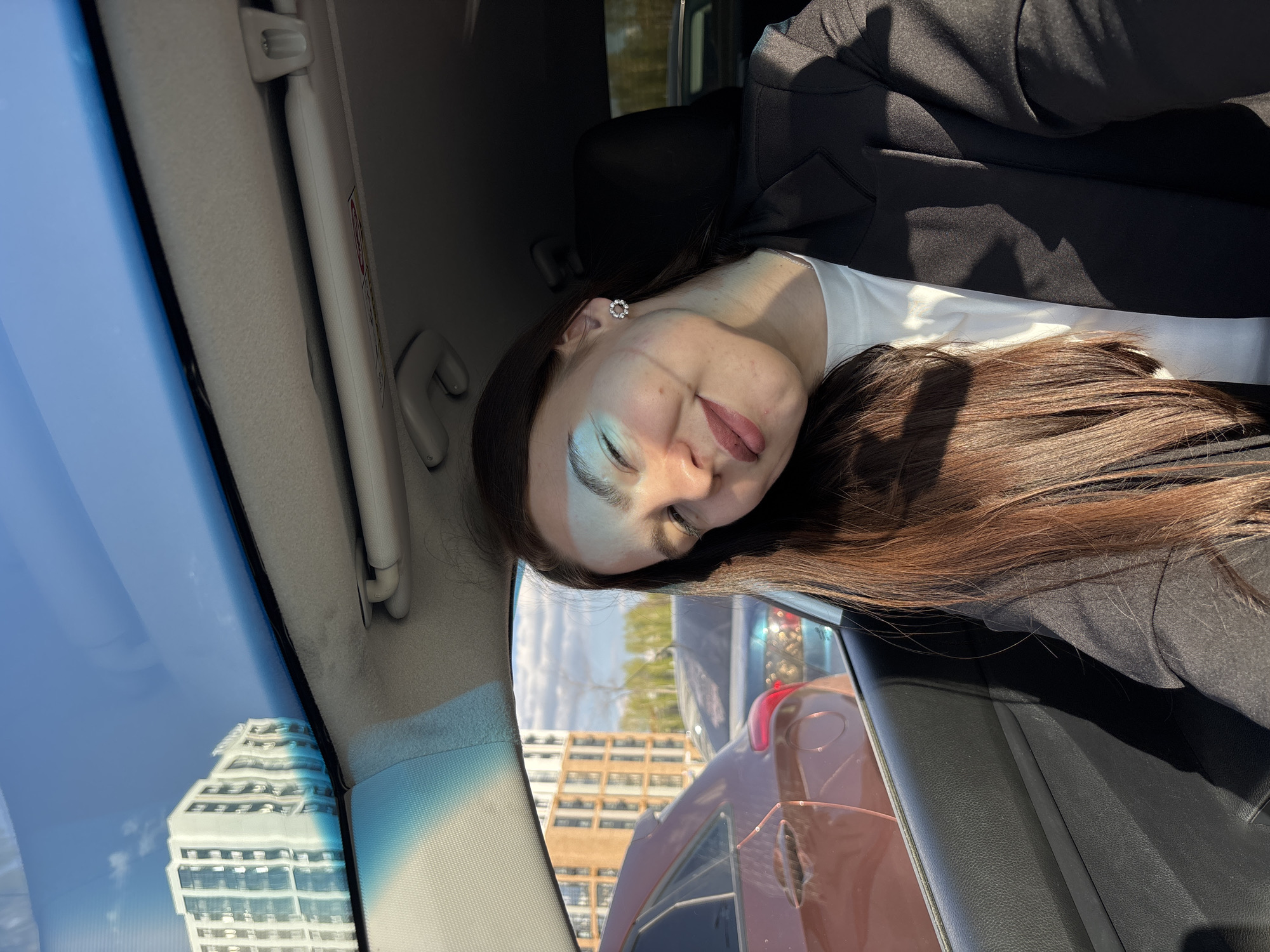 A young woman with long, auburn hair, wearing a white top and a black blazer, is sitting in the driver's seat of a car. Her eyes are closed, and her head is tilted to the side. The reflection of a modern high-rise building can be seen in the car's windshield. Through the car window, a multi-story brick building and the rear of another vehicle are visible. The scene is likely taken in an urban environment, possibly Perm, Russia, as per the prompt.