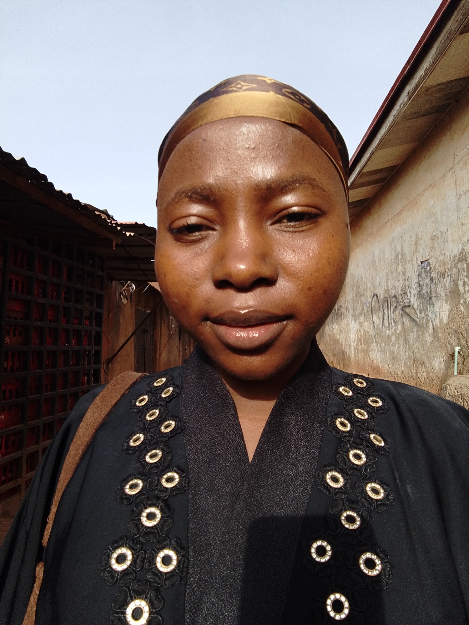 A close-up shot depicts a woman with brown skin, wearing a gold and brown head covering. She is looking directly at the camera with a slight smile. Her clothing is black with decorative floral patterns that have gold embellishments. She has a shoulder bag. The background includes a building with a textured wall covered in graffiti and another structure with red brick-like components. The sky is pale blue. Location: Unknown, Unknown.