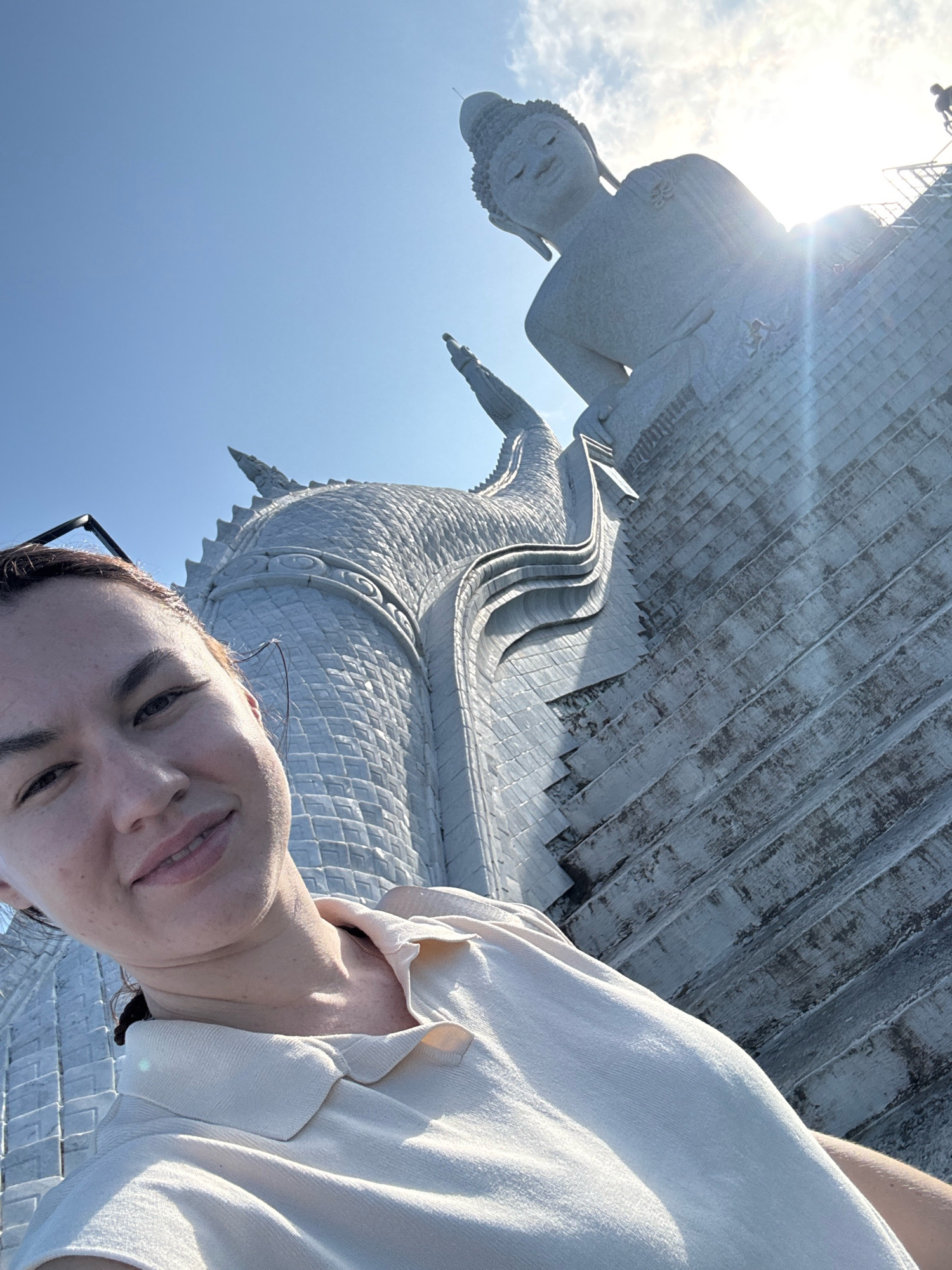 A low-angle selfie shows a woman in the foreground with a large, white Buddha statue in the background. The woman is smiling and looking at the camera. She is wearing a light-colored collared shirt. The Buddha statue is partially visible, with its head and torso in view. The statue is made of white stone and is adorned with intricate carvings. The background is a bright blue sky with some clouds. The scene appears to be in Karon, Thailand, specifically at the Big Buddha of Phuket, a prominent landmark.