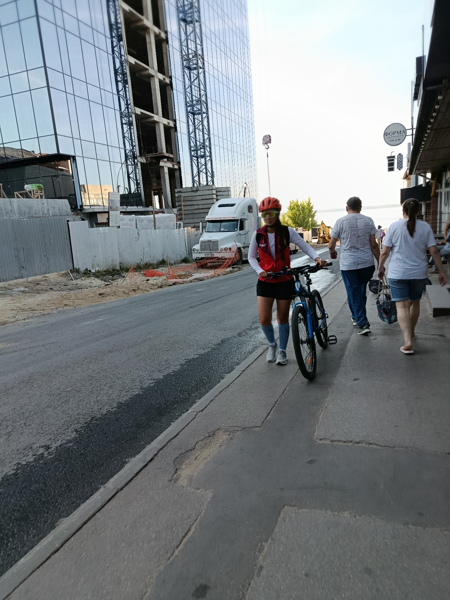 A female cyclist in an orange helmet and sunglasses walks her blue bicycle on a sidewalk next to a road. She wears a red vest over a white long-sleeved shirt and a black skirt. She is in the foreground, slightly to the left of center, with her back to the camera. Behind her, to the right, two people walk away from the camera on the sidewalk. The man wears a blue shirt and jeans, and the woman wears a white t-shirt and denim shorts, carrying a patterned bag. To the left of the road, a modern building with a glass facade is under construction, with a white truck and construction materials visible. The scene appears to be in a city, possibly along a waterfront, with a body of water visible in the distance. The overall lighting suggests it is daytime.
