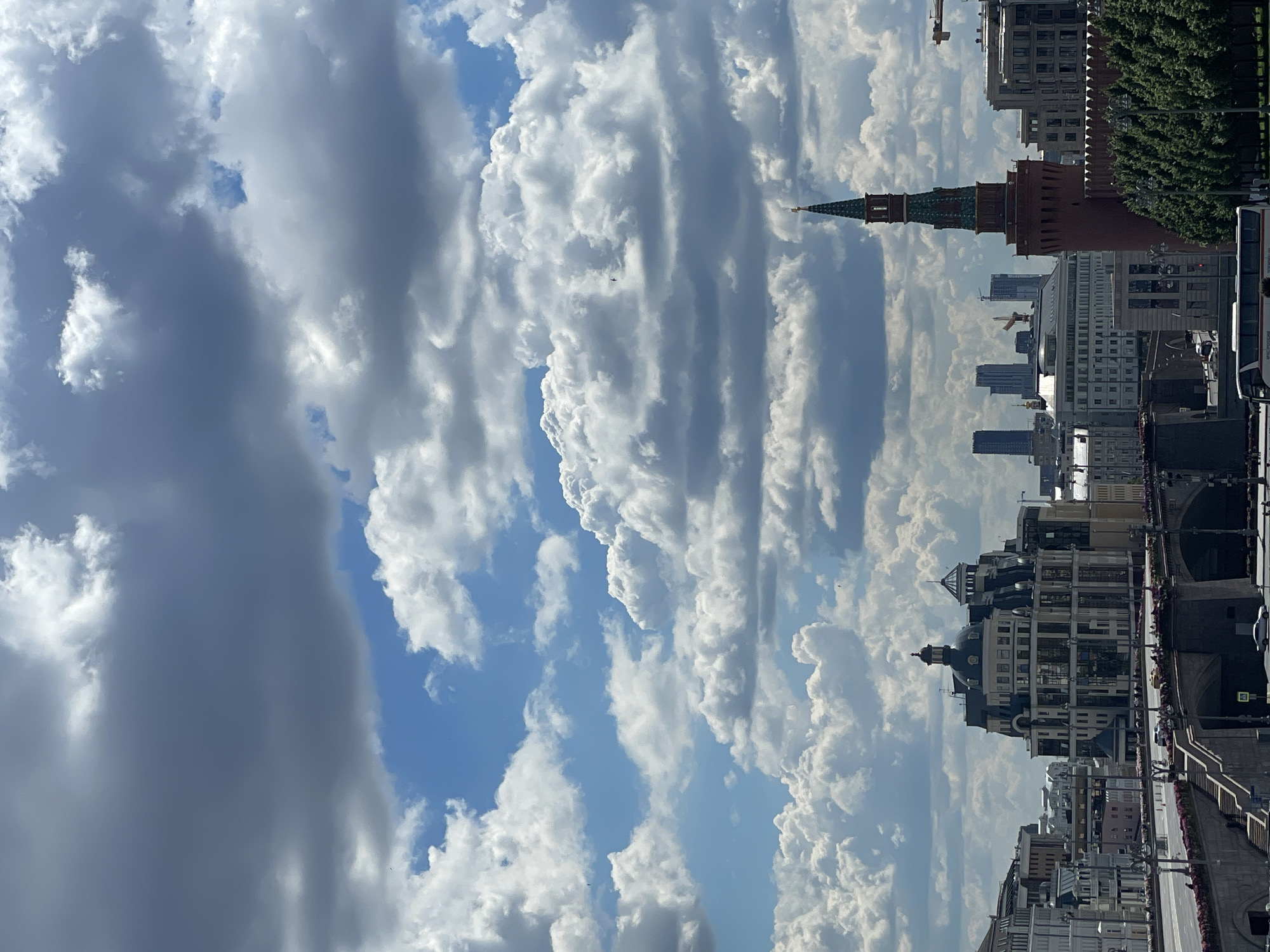 The image is a vertical, wide-angle shot of a city skyline under a cloudy sky. The dominant subjects are the numerous buildings, ranging in architectural styles from historic to modern, and the dramatic cloud formations. The clouds are a mix of dark, ominous storm clouds and bright, fluffy cumulus clouds, creating a dynamic contrast against the blue sky.
The scene appears to be a daytime shot, likely late morning or early afternoon, given the bright illumination on the buildings and the intensity of the clouds. The weather seems to be changing or mixed, with both sunny and stormy elements present in the sky.
Notable details include a tall, spired tower, possibly a clock tower or part of a historic structure, which stands out among the other buildings. There are also cranes visible in the background, suggesting ongoing construction or development in the city. The perspective is from a slightly elevated viewpoint, looking down and across the cityscape. The presence of a bus on the right side of the frame indicates a public thoroughfare or street.
No people are clearly identifiable in the image, and no specific activity or event is taking place; it's a static view of the urban landscape. There is no visible text within the image. The overall mood is one of grandeur and the power of nature juxtaposed with human construction, with the dramatic sky adding a sense of awe or perhaps foreboding. The rotation of the image suggests it was captured at an unusual angle or has been rotated post-capture.