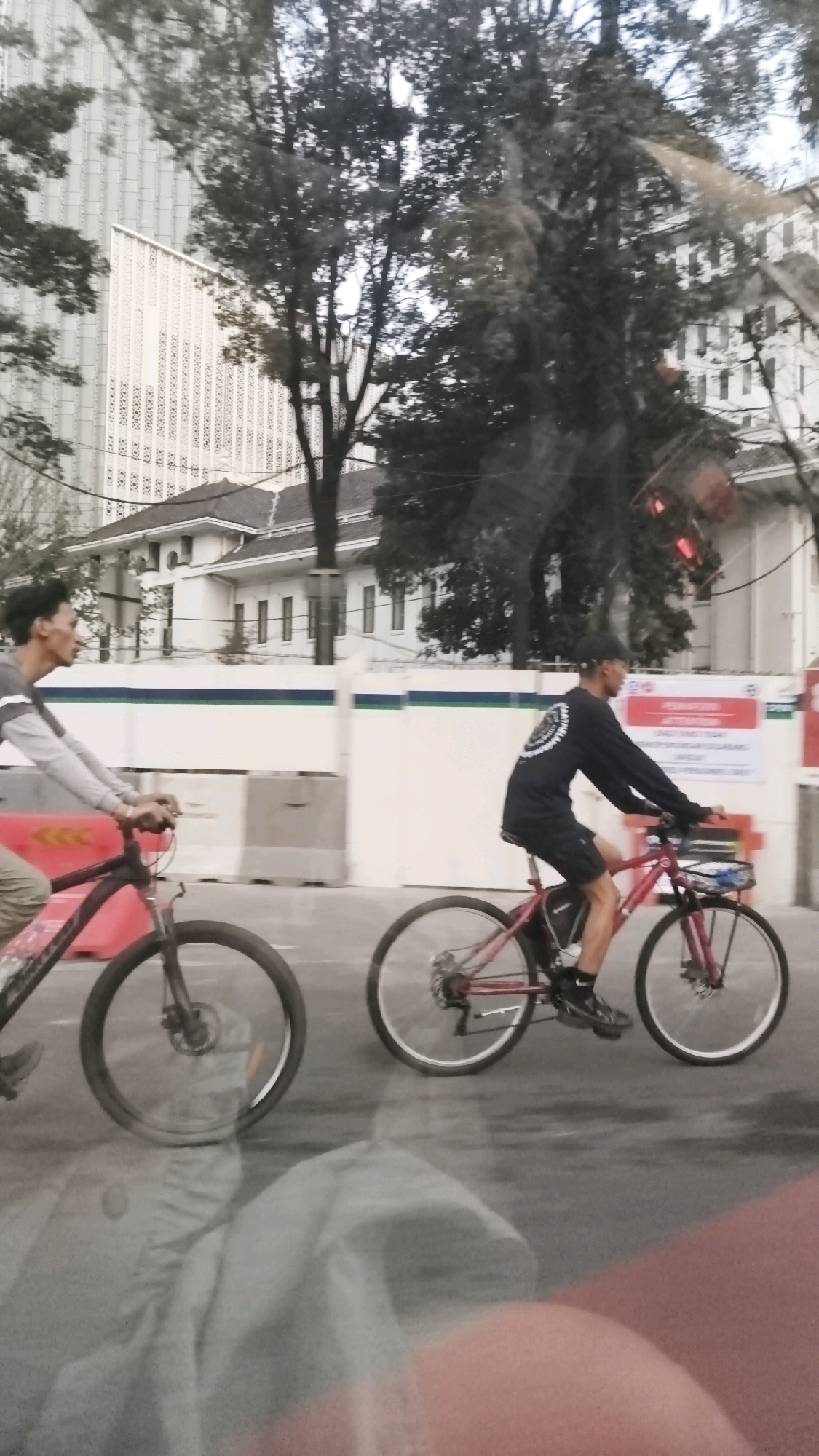 In Central Jakarta, Indonesia, two men are riding bicycles on a street, possibly during daylight hours. The man in the foreground, on the left, is wearing a grey long-sleeved shirt with white stripes on the sleeves and light-colored pants. He is riding a black bicycle with a red front frame detail. The second man, on the right, is wearing a black long-sleeved shirt with a circular graphic on the back and black shorts. He is riding a red bicycle. Both bicycles appear to have front baskets. They are riding on a paved road, with a white barrier and greenery visible in the background, along with buildings that suggest an urban environment. The image has a reflective quality, indicating it might have been taken from inside a vehicle.