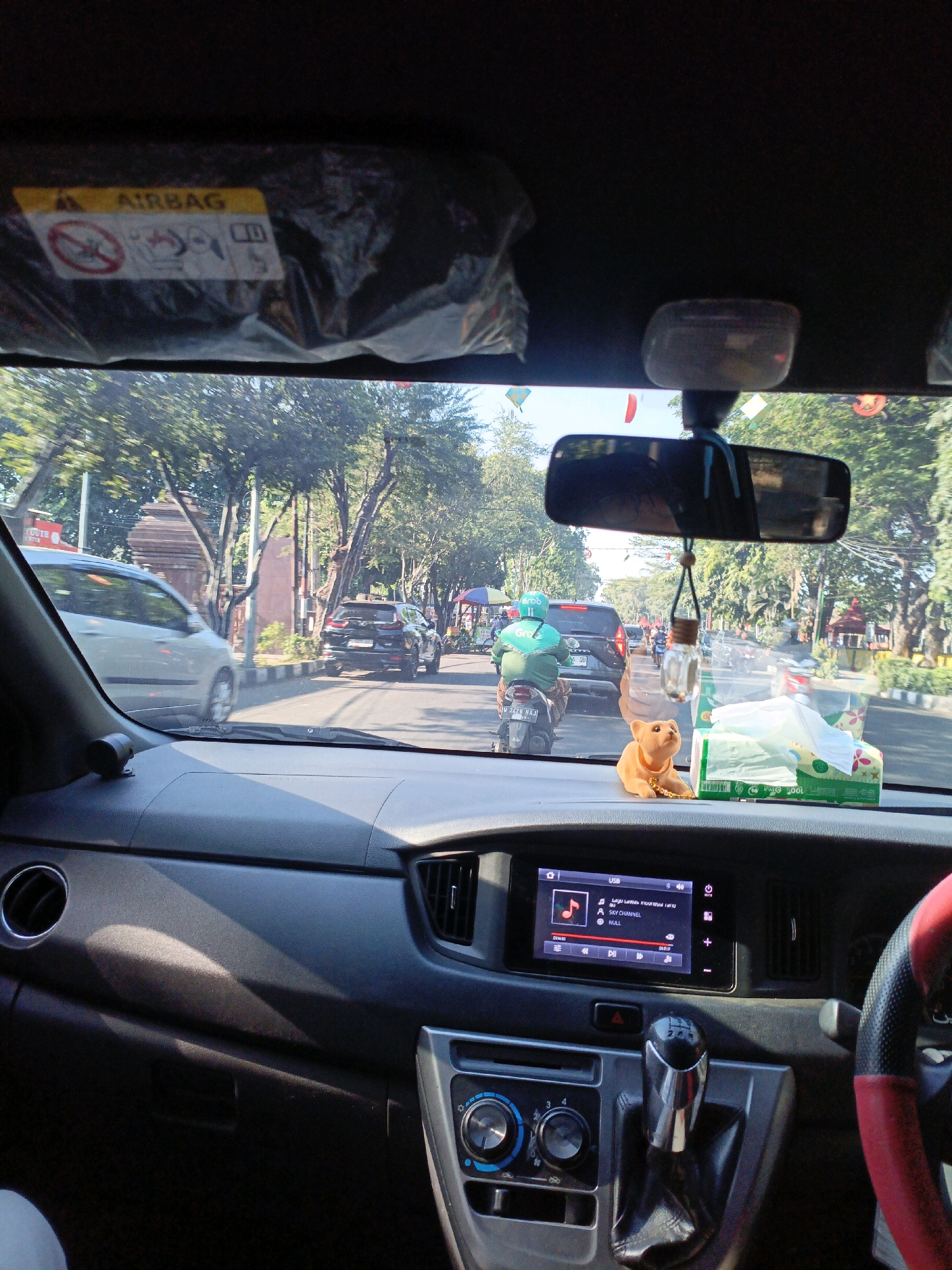 The image is taken from inside a car in Sidoarjo, Indonesia, looking forward through the windshield. The dashboard is visible in the foreground, with a touchscreen infotainment system displaying a music player, air conditioning controls, and a gear shifter. A small orange figurine of a dog sits on the dashboard next to a box of tissues. Outside the car, a street scene unfolds with trees lining the road. Several vehicles, including cars and a motorcycle with a rider wearing a green jacket, are present on the road. The weather appears to be sunny, casting shadows on the dashboard. An airbag warning sticker is visible on the sun visor.