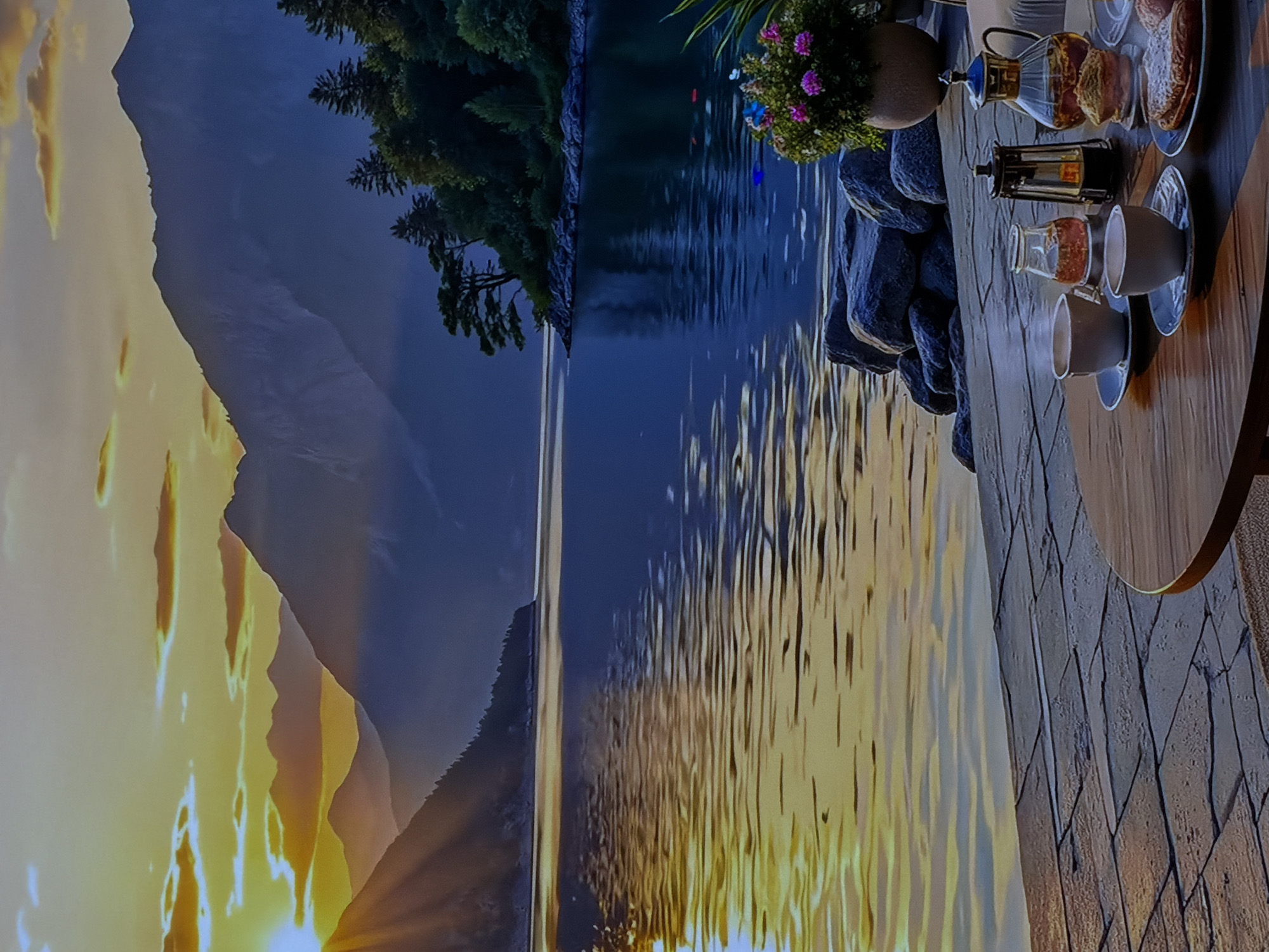 The image, appearing to be rotated 90 degrees clockwise, presents a serene outdoor scene at either sunrise or sunset, overlooking a vast body of water and mountains. In the immediate foreground, a wooden deck or pier features a round wooden table set for a meal or drink. On the table, there are two white ceramic cups with saucers, a clear glass bottle containing a reddish liquid, a glass carafe with a dark beverage, a dark French press coffee maker, and a plate piled with pastries or bread. Adjacent to the table, a potted plant with pink/purple flowers and green foliage rests on a decorative pile of dark grey rocks.
The midground is dominated by a large body of water, reflecting the golden light of the low sun in a shimmering pattern. The far shore is lined with dense evergreen trees that climb the lower slopes of majestic mountains. The sky above the mountains displays a gradient from bright golden hues near the horizon to deeper blues and grays, with some warm-toned clouds. The time of day is clearly dawn or dusk, creating a tranquil and picturesque ambiance. No people are visible, but the setup suggests an inviting moment of relaxation amidst this stunning natural backdrop, possibly in Sofiivska Borshchahivka, Ukraine. There is no visible text.