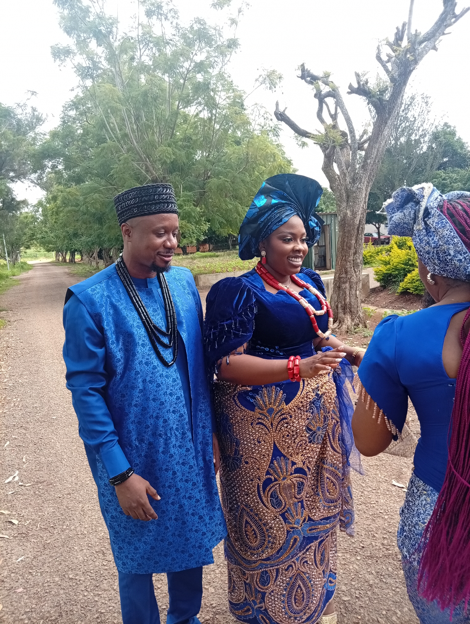 In Tambes Mile Four, Nigeria, three individuals are posing for a photograph outdoors on a dirt road lined with trees and vegetation. A man on the left, dressed in a blue traditional Nigerian attire and a black beaded cap, smiles as he looks down. He is wearing multiple strands of black beaded necklaces. To his right, a woman, adorned in a vibrant blue velvet top with intricate gold embroidery and a matching headwrap, engages in conversation. She is wearing large red bead necklaces and bracelets. Facing away from the camera, a third individual on the far right is partially visible, wearing a blue garment and a blue headwrap with visible magenta braids. The background features lush green trees and a clear sky, suggesting a daytime setting.