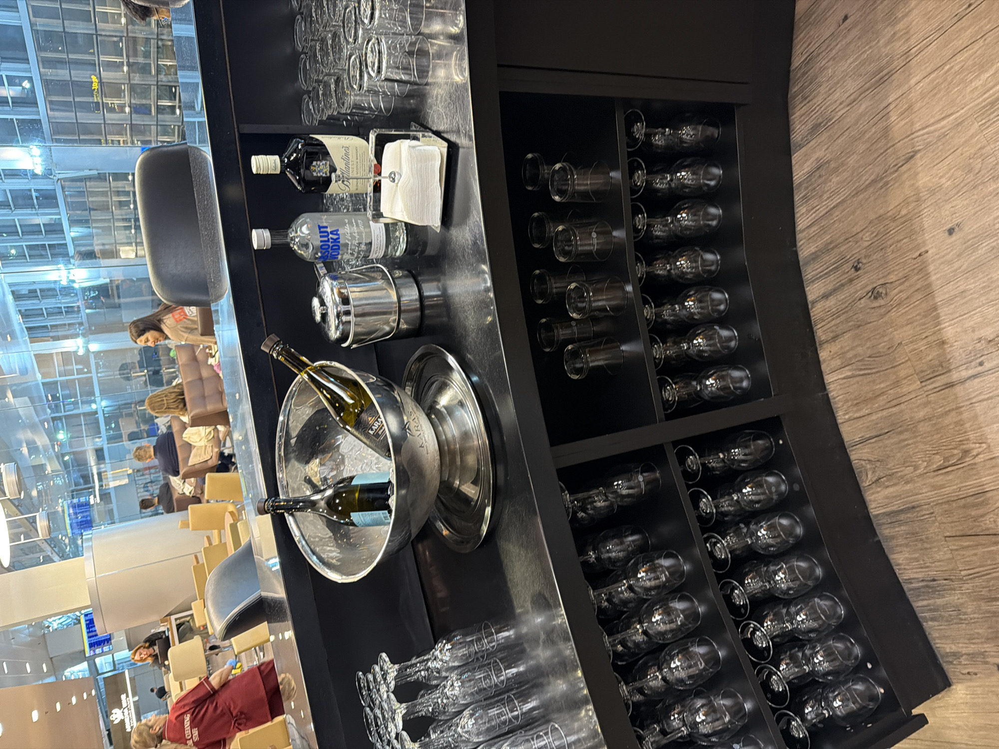 The image captures a bar area in what appears to be the Zvartnots International Airport lounge in Yerevan, Armenia. The bar features a black counter and curved shelving unit. The shelves contain an array of glassware including wine glasses, tall glasses, and shot glasses. A stainless steel ice bucket contains two bottles of white wine. On the counter are two liquor bottles, one labeled "Ballantine's" and the other "Absolut Vodka," as well as a stack of white napkins. In the background, lounge seating is visible along with people sitting, relaxing, or waiting. The location is within a building with large windows offering a view of the airport exterior and possibly the city.