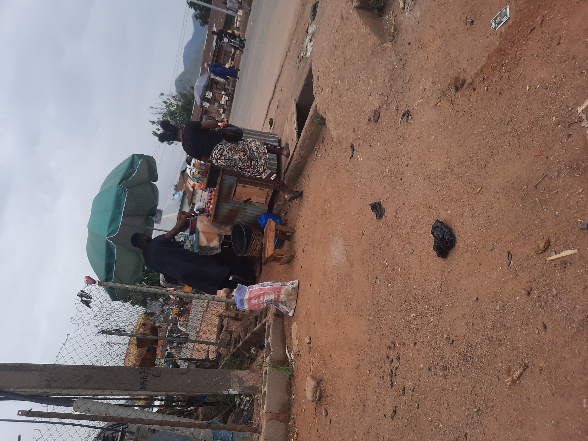 The image depicts a street scene in Gwong, Nigeria. A woman is buying food from a street vendor. The vendor is standing behind a makeshift stand with an umbrella. A black trash bag and litter are visible on the unpaved road. A chain-link fence and concrete structure border the scene on one side. In the background, there are buildings and a mountain range under a grey sky.