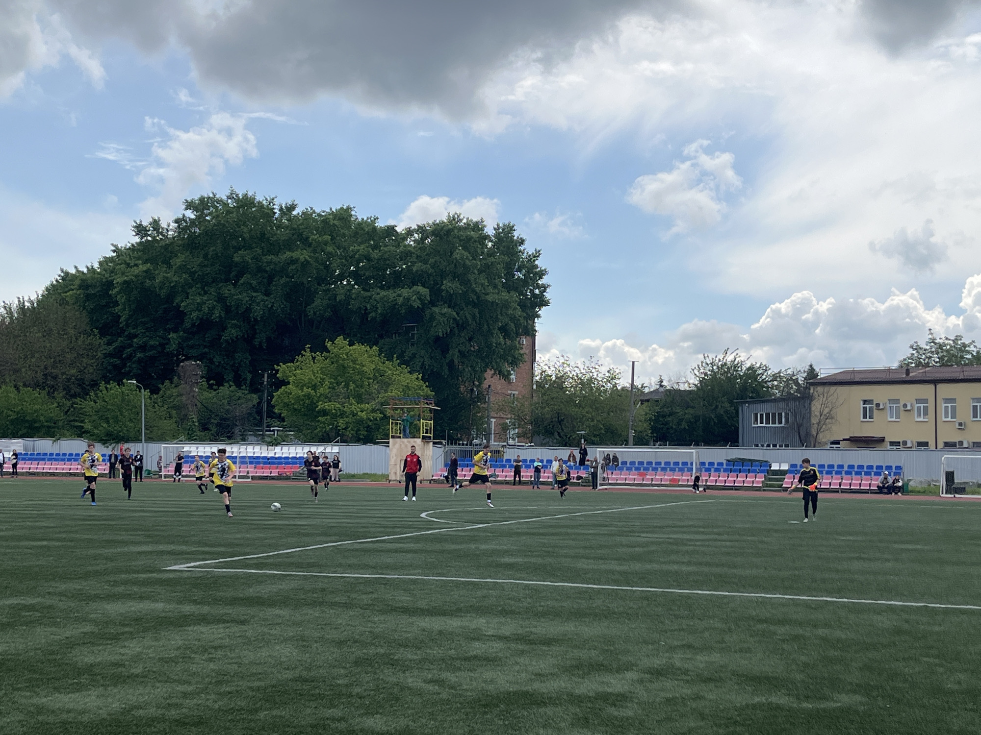 A soccer match is in progress on a green field. The field has white lines marking its boundaries. Players in black uniforms and yellow uniforms are actively engaged in the game. An adult, wearing a red jacket, is overseeing the match. In the background are rows of multi-colored stadium seats. Trees surround the edges of the playing field, along with some buildings. The sky is partly cloudy. The soccer field is located in the city of Krasnodar, Russia.