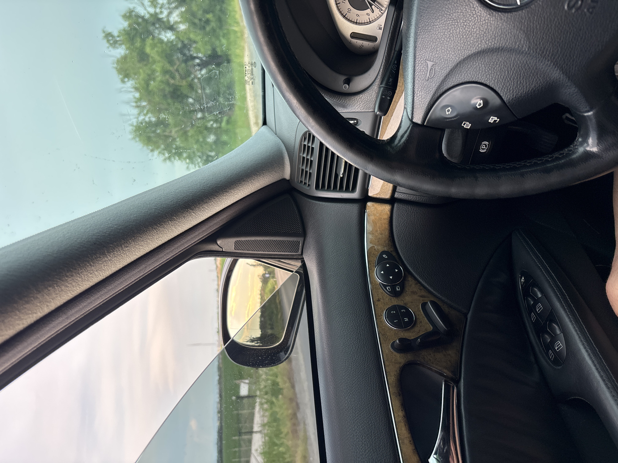 The image is a first-person perspective from inside a car, looking out the driver's side window and towards the steering wheel. The car appears to be parked on the side of a rural road in Yoakim Gruevo, Bulgaria. The interior of the car shows black leather upholstery on the door panel, wood trim accents, and a steering wheel with various controls. The window is slightly down, revealing a hazy sky with soft, warm light suggesting either sunrise or sunset, casting a golden reflection in the side-view mirror. Outside, there's a green landscape with fields and trees, bordered by a fence and a road. The overall impression is one of a peaceful, rural setting.