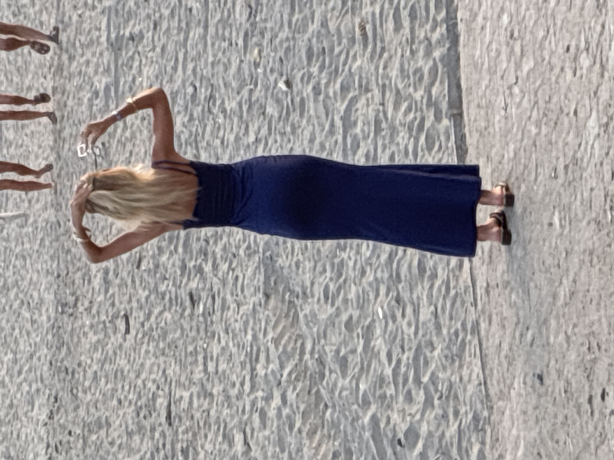 A woman with blonde hair, wearing a dark blue maxi dress and brown sandals, stands on a sandy beach. She is holding a pair of glasses in her right hand and has her left hand on her head. In the background, several people's legs are visible walking on the sand. The scene appears to be on a beach in Alanya, Turkey.