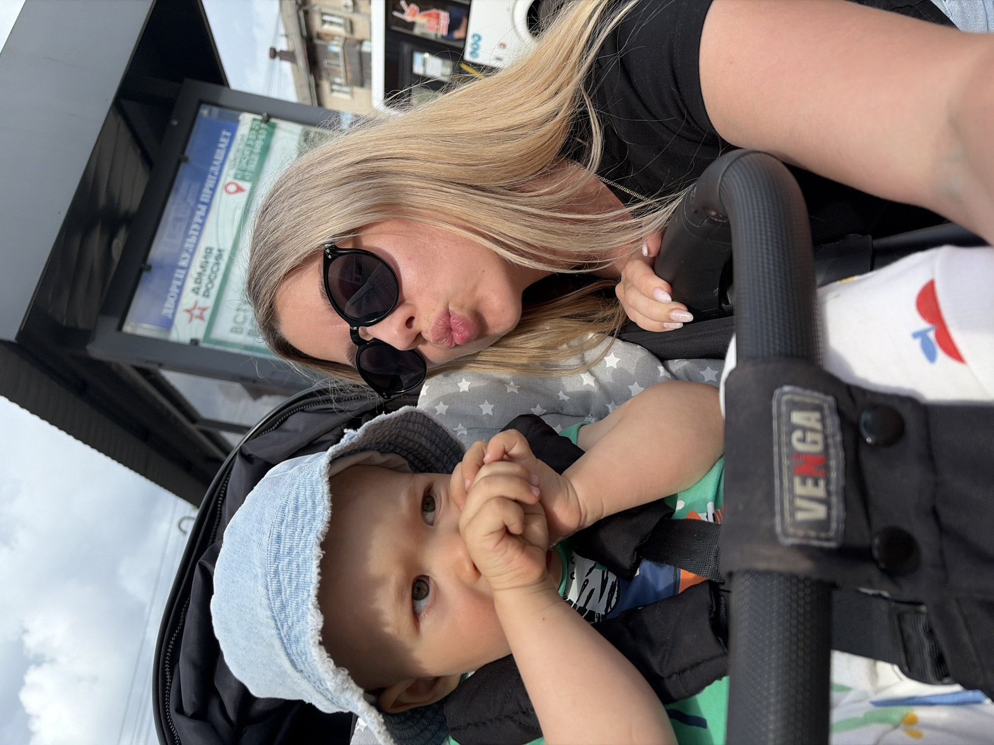 An outdoor medium shot captures a young woman and a baby in a stroller on a cloudy day. The woman, with blonde hair and black sunglasses, leans towards the baby, puckering her lips as if for a kiss. She wears a black t-shirt and has her hand resting on the stroller's handlebar. The baby, wearing a light blue denim hat and a green shirt, is looking to the side with its hands near its mouth. The stroller has a black frame and is partly visible. In the background, a bus stop sign with Russian text and images is mounted on a black structure. Beyond the bus stop, buildings and a white bus are visible. The scene appears to be in a city, possibly Chaikovsky, Russia, given the Russian text on the sign.