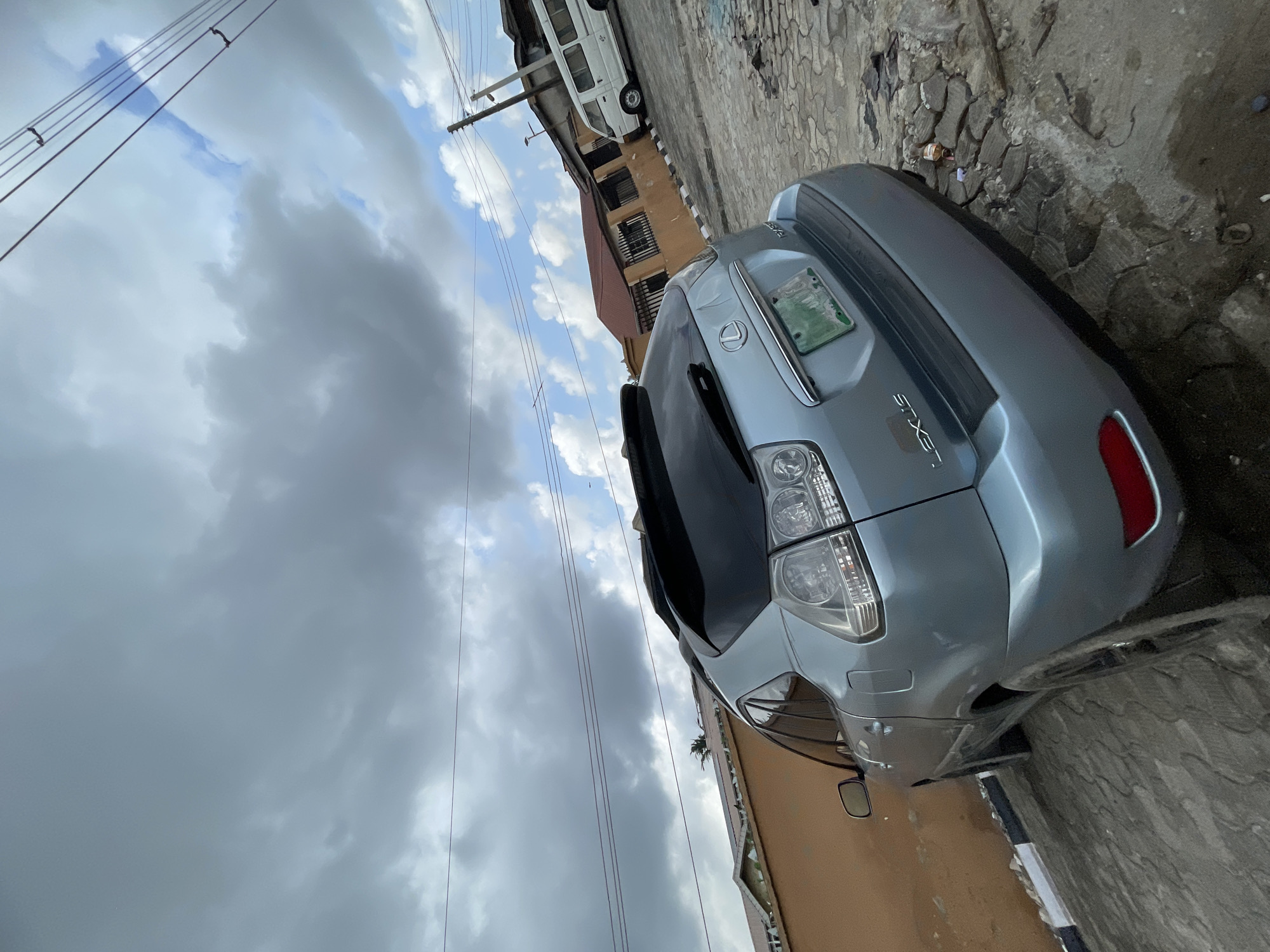This image, rotated counter-clockwise, presents an outdoor street scene with vehicles, buildings, and a cloudy sky. The main subject is a light grey Lexus SUV, seen from its rear and part of its side, parked on the right side of the frame. The word "LEXUS" is visible on the tailgate, and a green license plate is affixed, though its specific characters are unreadable.
The ground comprises a mix of interlocking concrete blocks and rougher, possibly broken, pavement or dirt. Near the rear of the Lexus, some litter, including a plastic bottle, is visible on the ground.
In the background, a light brown or tan building with dark-barred balconies stands behind the Lexus. Further along the street, a white minibus or commercial van is parked near a utility pole, partially obscured by other structures.
The sky is heavily overcast with prominent dark grey clouds, but patches of brighter blue sky are also visible. Multiple utility lines or power cables crisscross the upper portion of the frame. The lighting suggests it is daytime, under diffused light conditions. No people are visible in the scene. The overall setting appears to be an urban or semi-urban area, consistent with the location context of Ado, Nigeria.