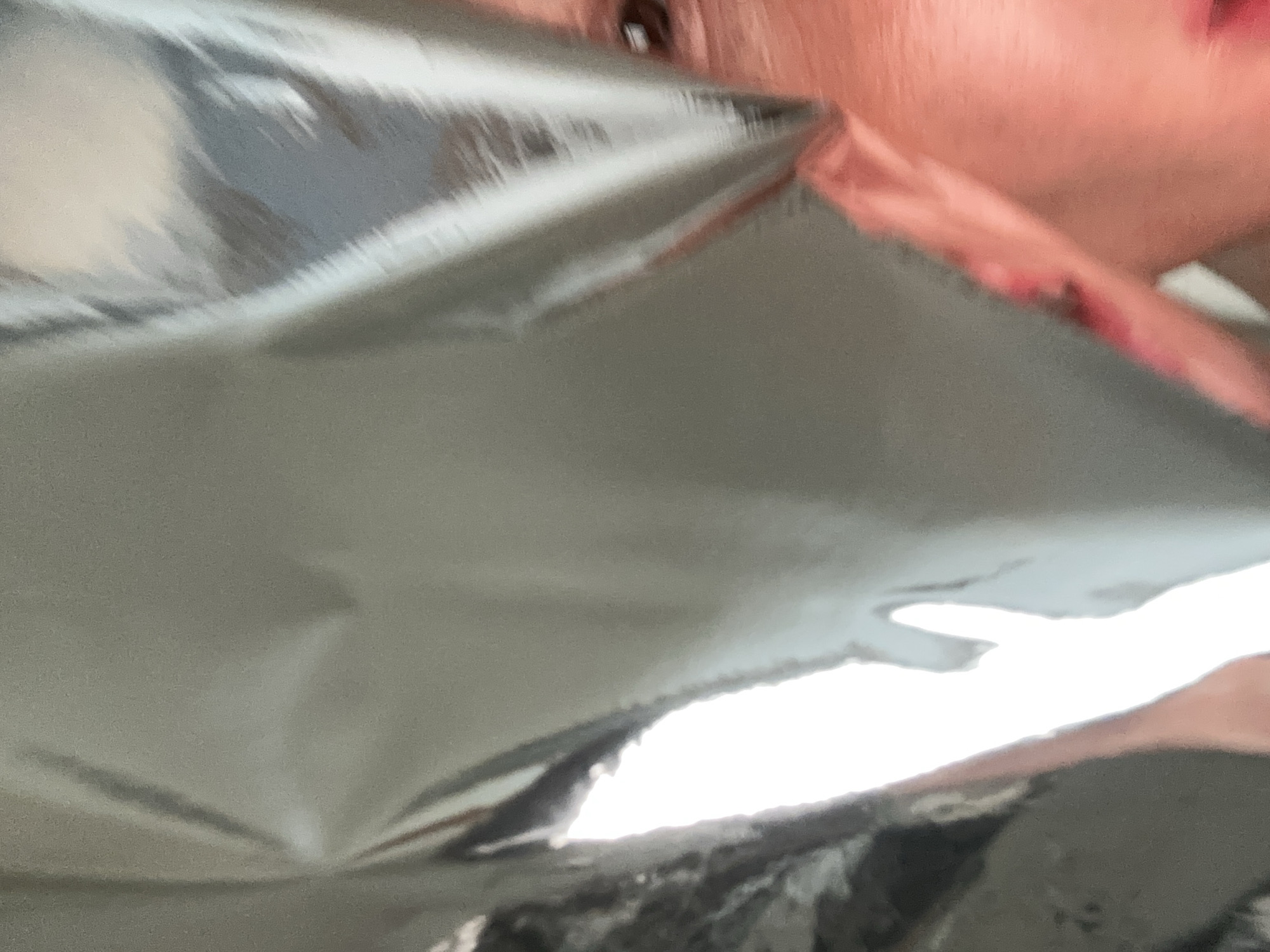 The image displays a close-up, highly reflective surface, possibly Mylar or metallic foil, dominating the upper and central portions of the frame with a metallic grey sheen and visible creases. A bright white light source is visible through an irregular opening or fold in this material on the left side. The lower right quadrant reveals a partial human face, specifically the right eye, eyebrow, and surrounding skin, which exhibits a reddish-pink tone. The iris is dark brown. No additional subjects, objects, or environmental details are discernible to establish a specific location within Tunbridge Wells, United Kingdom, or any other city.