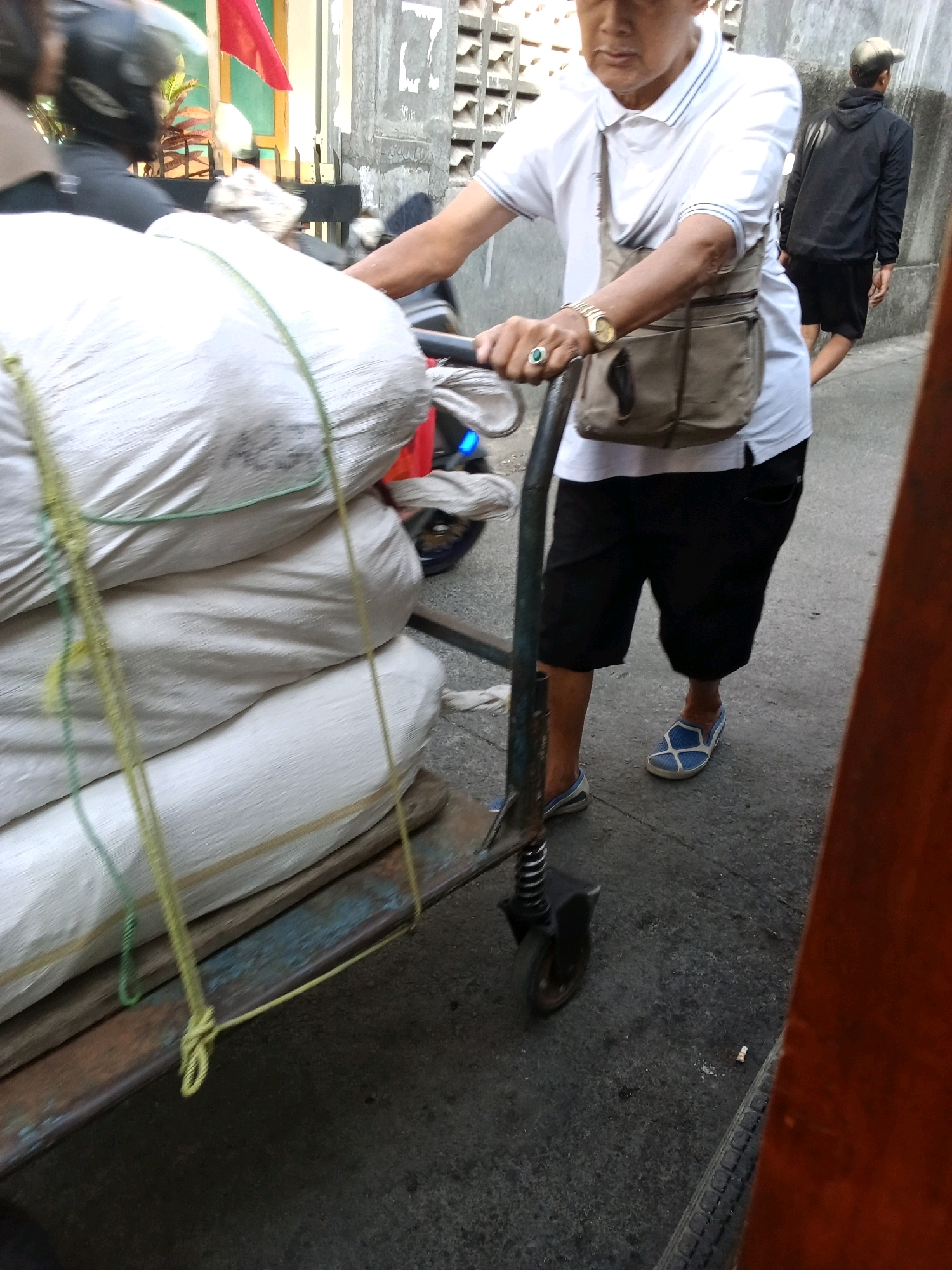 The image depicts a street scene in Bandung City, Indonesia. In the foreground, a man wearing a white polo shirt, black shorts, and blue and white slip-on shoes is pushing a cart loaded with large, white sacks. The sacks are tied with green and yellow ropes. The man has a beige messenger bag slung across his chest and is wearing a watch on his left wrist. He is holding onto the handlebars of the cart, which has a black base and a small wheel. Behind him, another man wearing a black hoodie and shorts walks away from the camera. The environment appears to be an urban street with a concrete wall to the left, possibly of a building. A red flag is visible on a pole in the background. The ground is paved with asphalt and shows some debris. The lighting suggests it is daytime.