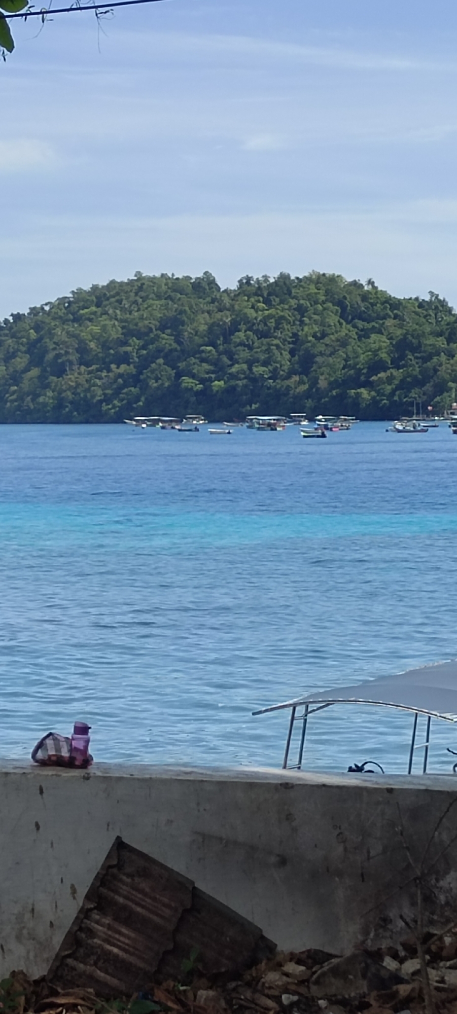 The image depicts a serene coastal scene in Sabang, Indonesia. In the foreground, a concrete wall runs horizontally, with a few discarded pieces of corrugated metal and dry leaves scattered below it. On top of the wall, near the left edge, a patterned bag and a purple water bottle are placed. Beyond the wall, a calm expanse of blue water stretches out, with a lighter turquoise band of shallower water visible in the mid-ground. A cluster of small boats with canopies are dotted across the water, suggesting a leisurely day. In the distance, a lush green, tree-covered island forms the backdrop. The sky above is a clear, pale blue with a few wispy clouds. The overall impression is one of tranquility and natural beauty.