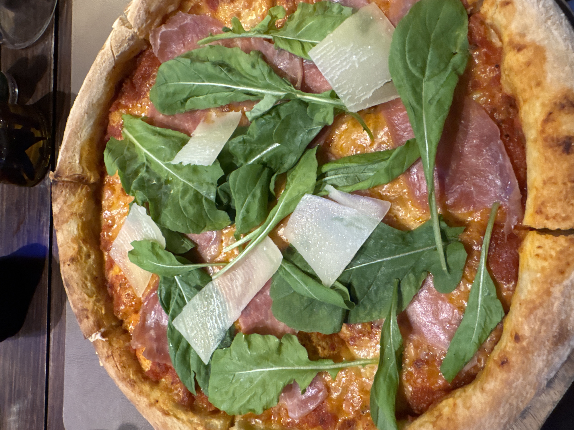 A top-down view reveals a pizza on a wooden surface. The pizza has a golden-brown crust and is topped with tomato sauce, prosciutto, arugula, and parmesan cheese shavings. A small bottle is visible in the upper left corner. The location is Baan Chaweng, Thailand.