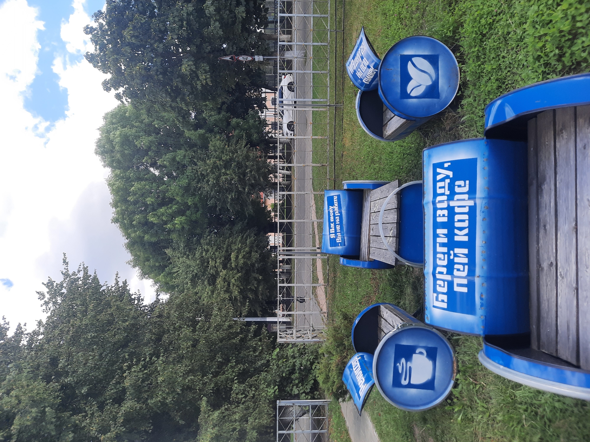 In Shklov, Belarus, an outdoor seating area is comprised of repurposed blue barrels cut in half to form benches. Three of these barrel benches are visible. The bench in the foreground is partially obscured by its own construction, with a wooden slat seat and blue barrel sides. To the left, another barrel bench is angled, showing a white emblem of a steaming coffee cup on its side. To the right, a third barrel bench faces the viewer, featuring a white emblem of two coffee beans on its side. In the center, a barrel bench is positioned perpendicular to the others, with a wooden slat seat and a blue barrel body. Text in white Cyrillic is visible on the barrel sides. One portion reads "Береги воду, пей кофе" (Save water, drink coffee). Another portion, on the rightmost bench, is partially legible and seems to say "я Вас вижу - Вы не на работе" (I see you - you are not at work). The benches are situated on a grassy area. In the background, a white fence separates the grassy area from a paved road. A white modern car is driving on the road. Behind the road, a line of trees forms a dense green backdrop, with a cloudy sky overhead.