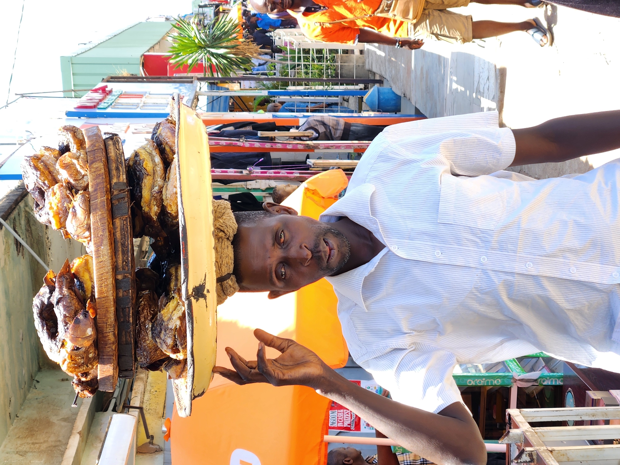 A man in Jos, Nigeria, balances a wooden tray laden with cooked fish on his head. He wears a light-colored striped shirt and extends his left hand. The tray is circular with a yellow rim and is stacked with several layers of grilled fish. A second man in an orange shirt and khaki shorts walks in the background. There are structures and foliage visible behind them, including parts of buildings and a palm tree.