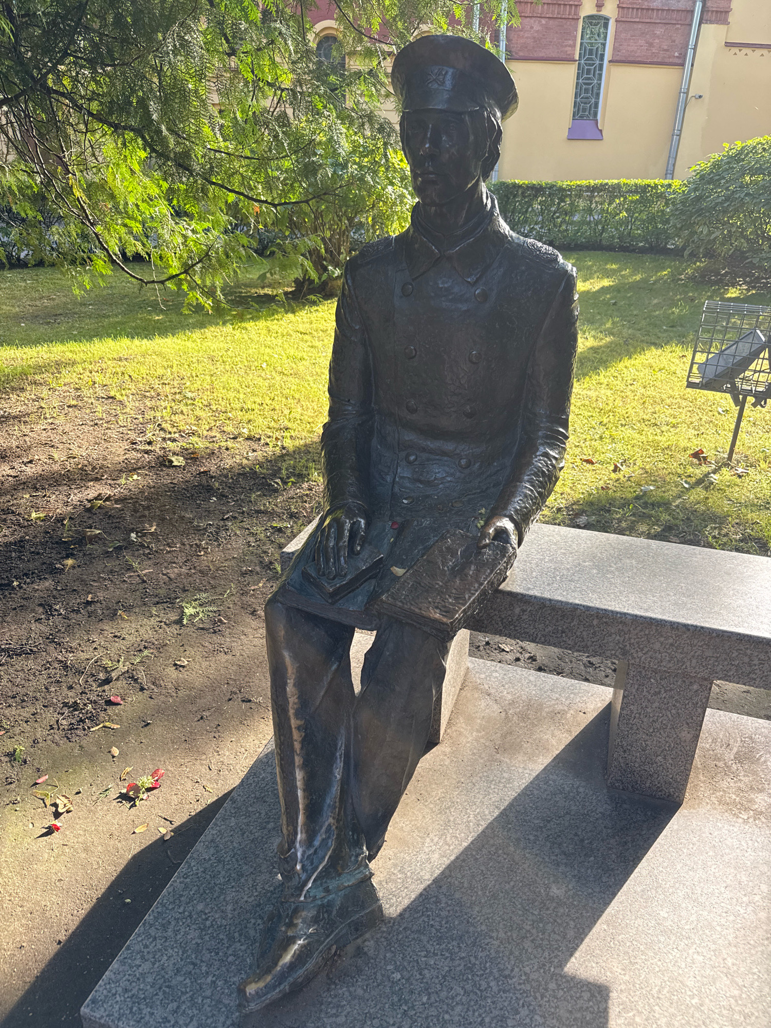 A bronze statue of a man in military uniform is seated on a granite bench in a park-like setting. The man is wearing a cap, a double-breasted coat with buttons, and trousers. He is holding an open book in his lap. The statue is in profile, facing to the viewer's left. The background includes a lush green lawn, trees with sunlit foliage, and a light yellow building with arched windows. There is a metal grate structure with a stone slab on a pole to the right of the bench. The ground in front of the bench is dark earth with scattered red leaves and fallen blossoms. The scene is illuminated by bright sunlight, casting long shadows. This statue is located in Saint Petersburg, Russia, specifically in the courtyard of the Former Officers' Assembly House on Liteyny Prospekt.