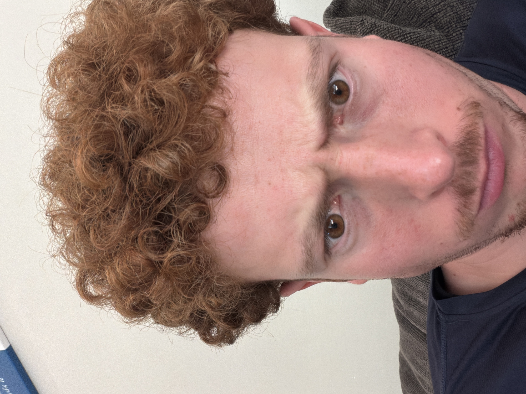 The original image is a close-up selfie of a person with curly red hair and brown eyes, looking directly at the camera with a furrowed brow. They are wearing a dark blue shirt. The background is a plain wall with a glimpse of a brown textured surface, possibly a couch. There are no discernible elements that would indicate the location in Pepper Pike, United States of America.