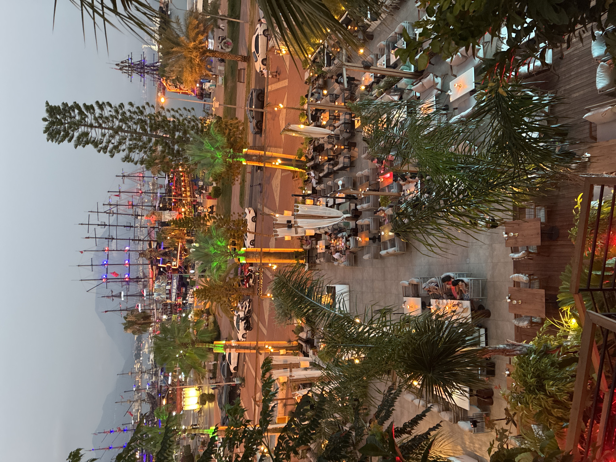 This is a high-angle, wide shot photograph taken at dusk in Alanya, Turkey, overlooking a waterfront promenade and an outdoor restaurant. In the background, several large, multi-masted sailing ships are docked, illuminated with colorful lights, some appearing to be pirate-themed. Mountains are visible in the far distance, shrouded in haze. The middle ground shows a street with parked cars and a sidewalk, lined with palm trees. The foreground and immediate middle ground are dominated by an outdoor restaurant area with numerous tables and chairs set up on a patio. People are seated at tables, dining and socializing. The restaurant is decorated with string lights and modern outdoor furnishings. Palm trees and other lush greenery are scattered throughout the scene, adding to the tropical ambiance. The overall atmosphere is lively and inviting, characteristic of a popular tourist destination at sunset.