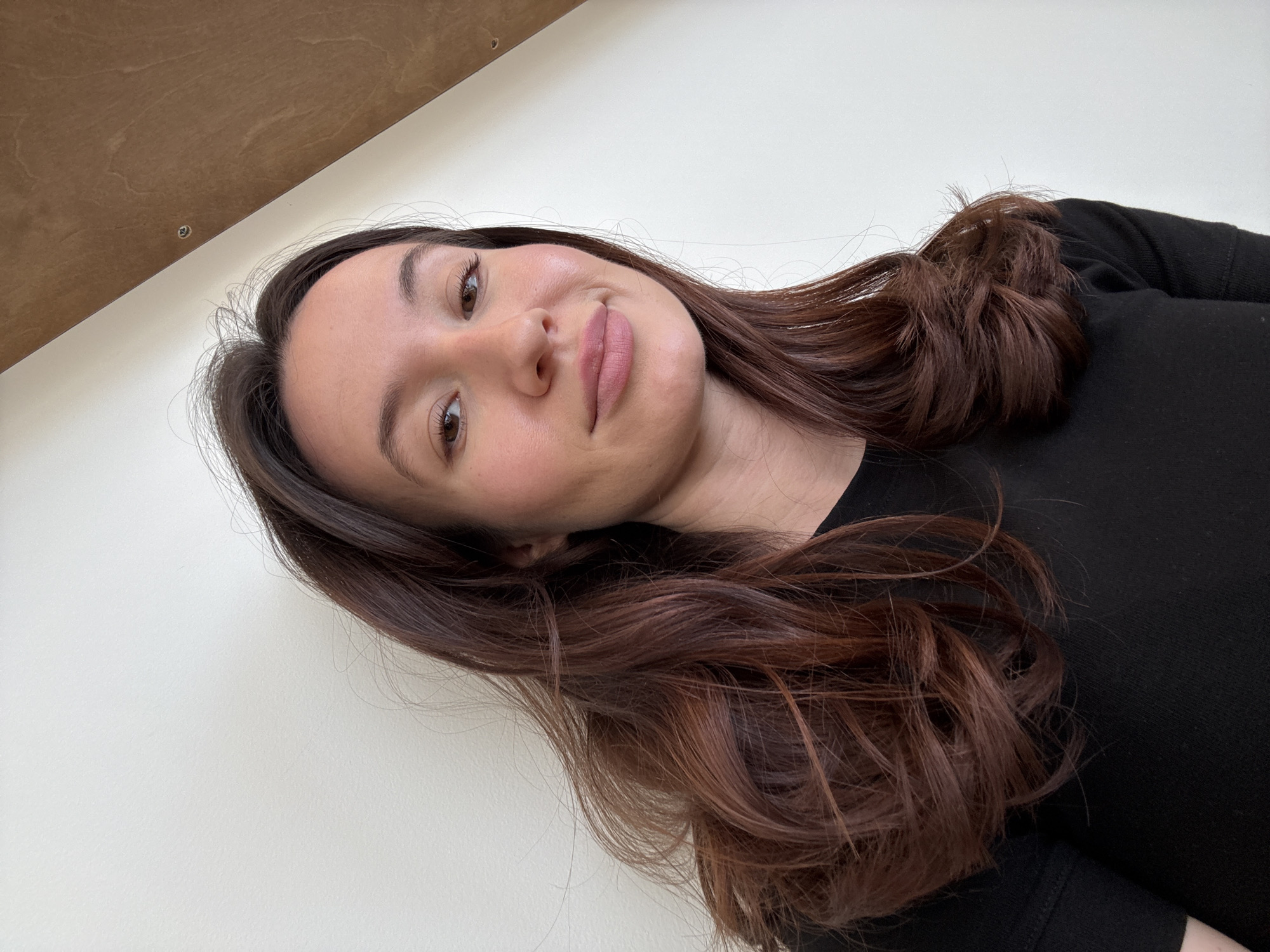 The image shows a woman with long, auburn hair, wearing a black shirt. She is looking at the camera with a slight smile. The background consists of a white wall and a section of a wooden panel. There is no information available in the image to suggest this photo was taken in Tel-Aviv, Israel or any other location.
