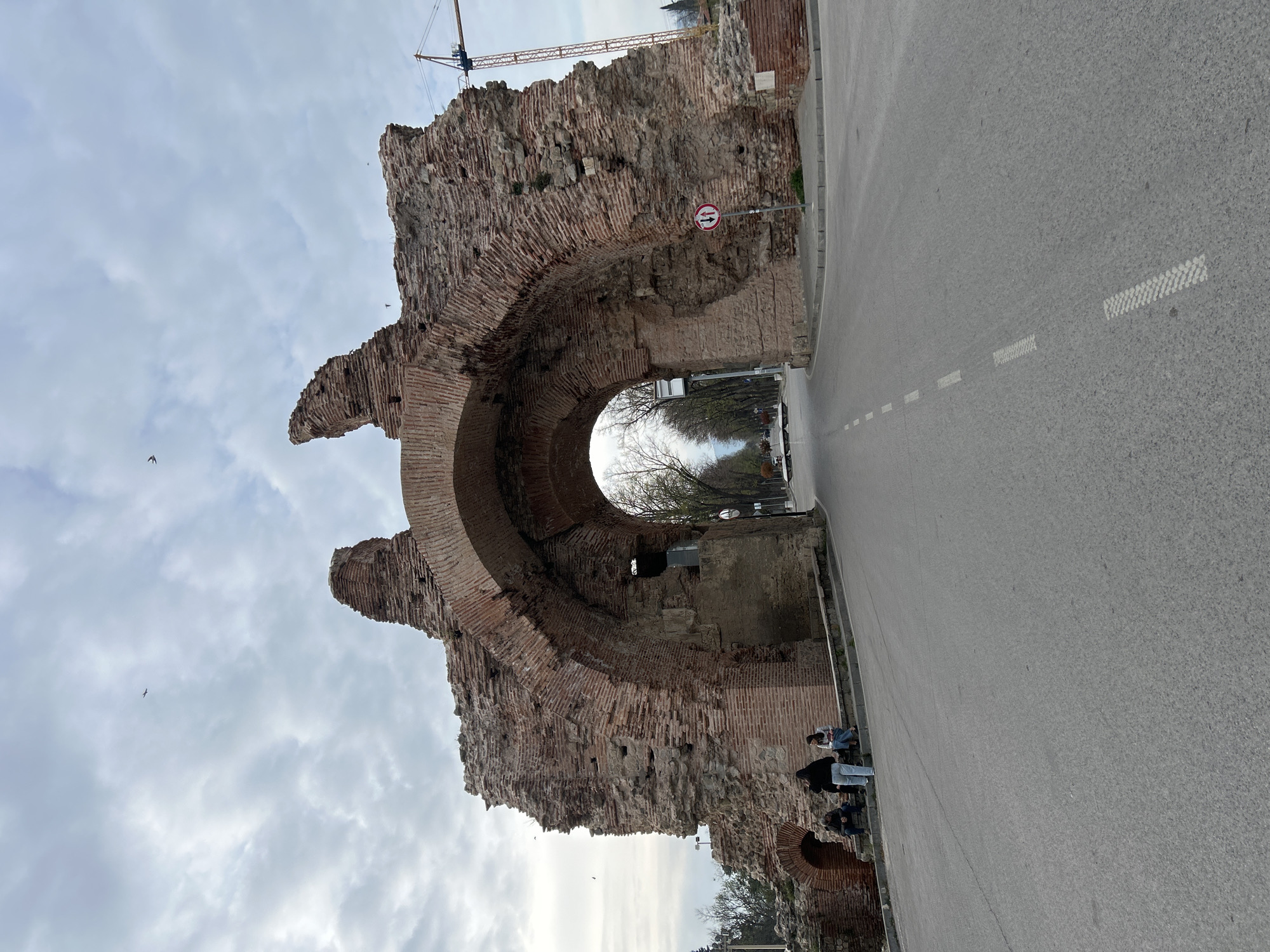 The image displays a significant portion of ancient brick and stone ruins, likely a Roman archway, under a cloudy sky in Hisarya, Bulgaria. The large arch, constructed from weathered reddish-brown bricks and mortar, dominates the left and central parts of the frame, showing its aged and partially collapsed structure. Through the arch's opening, a paved road extends into the background, flanked by trees with sparse or bare branches, suggesting either early spring or late autumn.
To the right of the archway, a modern asphalt road with dashed white lane markings runs parallel to the historical structure. Several people are visible near the base of the ruins on the right side of the arch. Some are seated on a low structure, possibly a bench, while others stand, seemingly observing the site. In the upper left background, behind the ruins, a large yellow construction crane is partially visible against the overcast sky. A circular red and white traffic sign, mounted on a pole near the archway, clearly displays two black arrows pointing horizontally in opposite directions, signifying two-way traffic. A few birds can be spotted flying in the cloudy sky. The overall scene captures a blend of ancient architecture and modern life on a visibly overcast day.