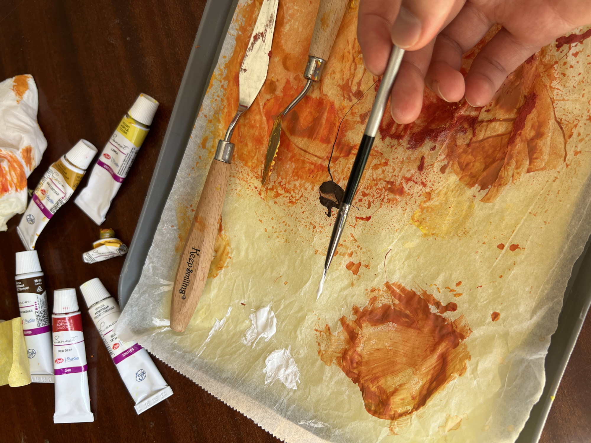 An overhead, eye-level shot shows an art studio with tubes of paint and a palette on a table. The scene is set on a wooden table. The art supplies are arranged on a baking tray lined with parchment paper. In the foreground, a hand holds a thin paintbrush loaded with brown paint over a palette of orange and yellow paint splatters on the parchment paper. Three palette knives are visible on the parchment paper, one with a wooden handle and a curved metal blade, and another with a wooden handle and a straight metal blade. Several tubes of paint are scattered in the background. The tubes are various colors including red, yellow, and brown, and some are partially squeezed. The overall impression is one of a messy, but productive, art creation process.