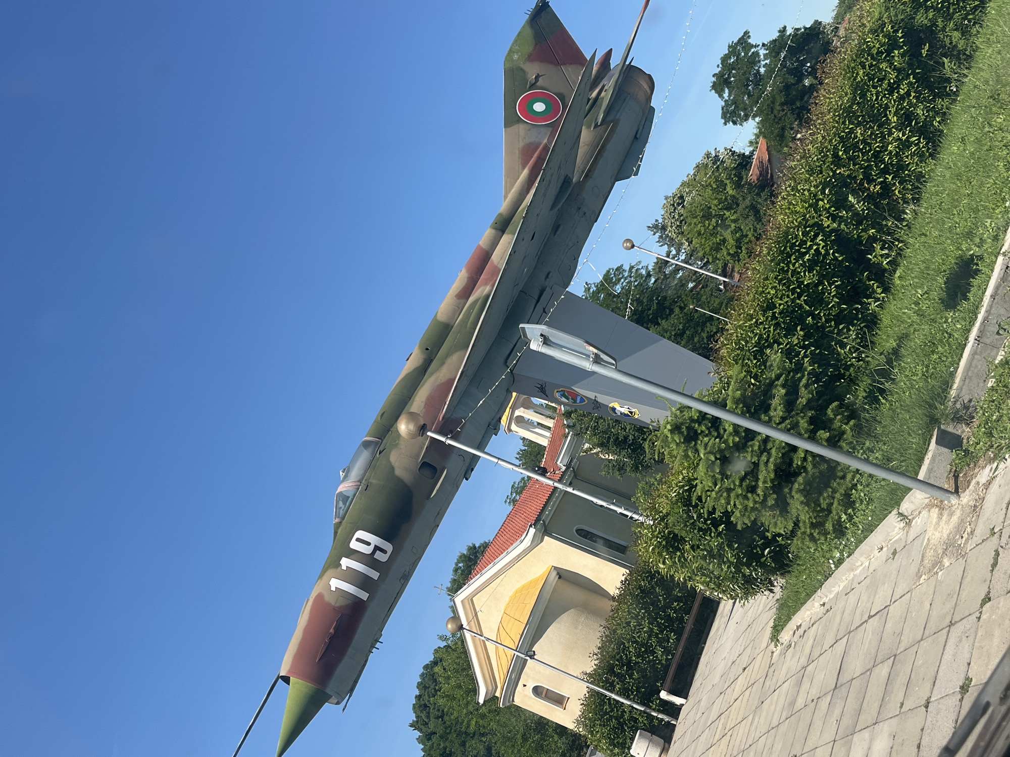 In the city of Ravnets, Bulgaria, a fighter jet, painted in camouflage green and brown with the number "119" prominently displayed on its fuselage, is mounted on a pedestal. The jet is angled upwards against a clear blue sky. To the left and below the jet, a small, yellow church with a red tile roof is visible. Lush green bushes and trees surround the church and the base of the jet's display. The foreground shows a paved walkway with some grass and weeds growing through the cracks. The overall scene depicts an outdoor monument or exhibit, likely in a public space.