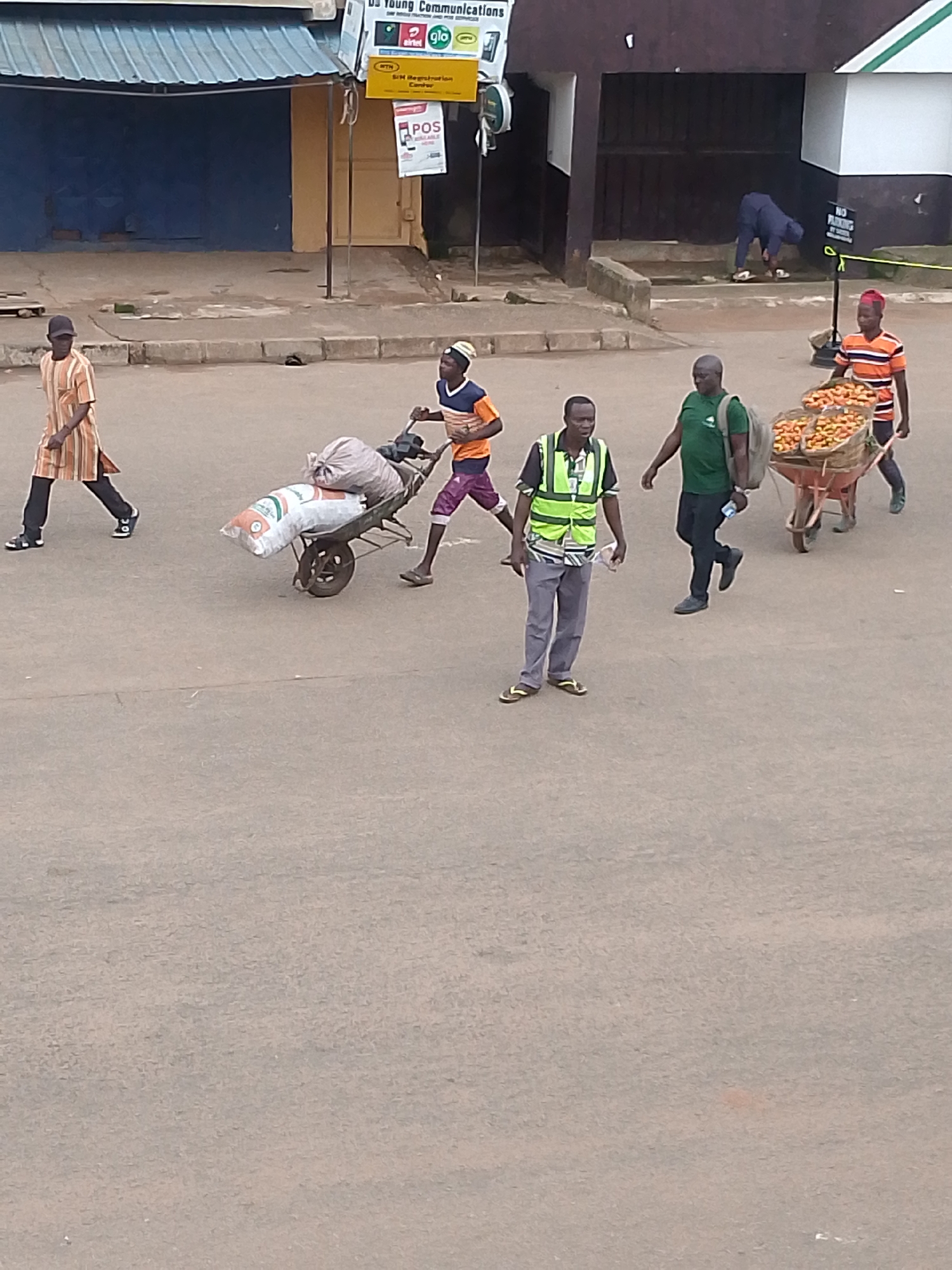 In Bukuru, Nigeria, on an overcast day, several individuals are engaged in street commerce and transit. A man in a high-visibility vest and grey pants stands in the foreground, holding a plastic bag. To his left, a person in an orange and white striped garment walks away from the camera. Behind him, two individuals are pushing a wheelbarrow laden with sacks, while another person walks ahead of them. To the right, a wheelbarrow filled with produce, possibly oranges, is being pushed by a person wearing an orange and white striped shirt. Further in the background, a person in blue clothing is crouched down, and a woman in an orange and white striped shirt is visible. The scene takes place on a paved road, with buildings and signage visible in the background, indicating a commercial area.
