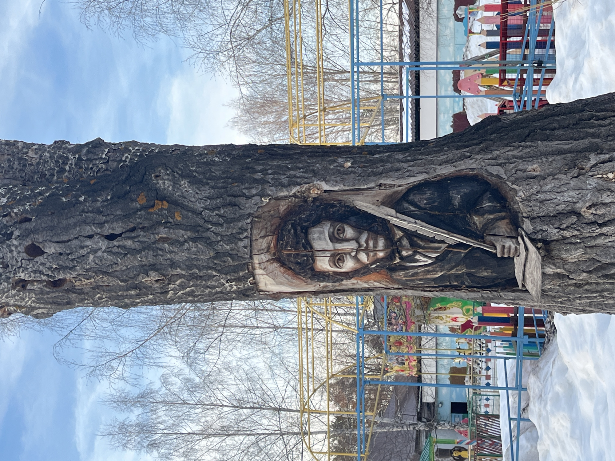 A large, dark-barked tree trunk stands prominently, featuring a detailed relief carving of a male portrait in a central recess. The carved figure, resembling Pyotr Ilyich Tchaikovsky, has dark hair, dark eyes, a mustache, and is depicted wearing a dark coat over a white shirt with a dark tie. His left hand, also carved in relief, holds a quill pen positioned over a flat surface, possibly a book or musical score. The tree bark around the carving is textured and dark, with several visible cavities higher on the trunk.
The ground surrounding the tree is covered with patches of snow. Behind the tree, a colorful children's playground or park area is visible. This area includes a blue metal fence and a yellow metal climbing or support structure. Brightly painted wooden cutouts shaped like oversized pencils in red, yellow, blue, pink, green, and white form a decorative barrier. Portions of an amusement ride with ornate gold, red, and blue decorations, possibly carousel horses or swing seats, are discernible. A large pink mushroom-shaped object is also present.
In the background, numerous leafless deciduous trees rise against a bright blue sky with scattered white clouds. Low-rise buildings with dark roofs and a structure with a blue facade are partially visible through the playground equipment. The scene depicts an outdoor winter environment in the city of Chaikovsky, Russia.