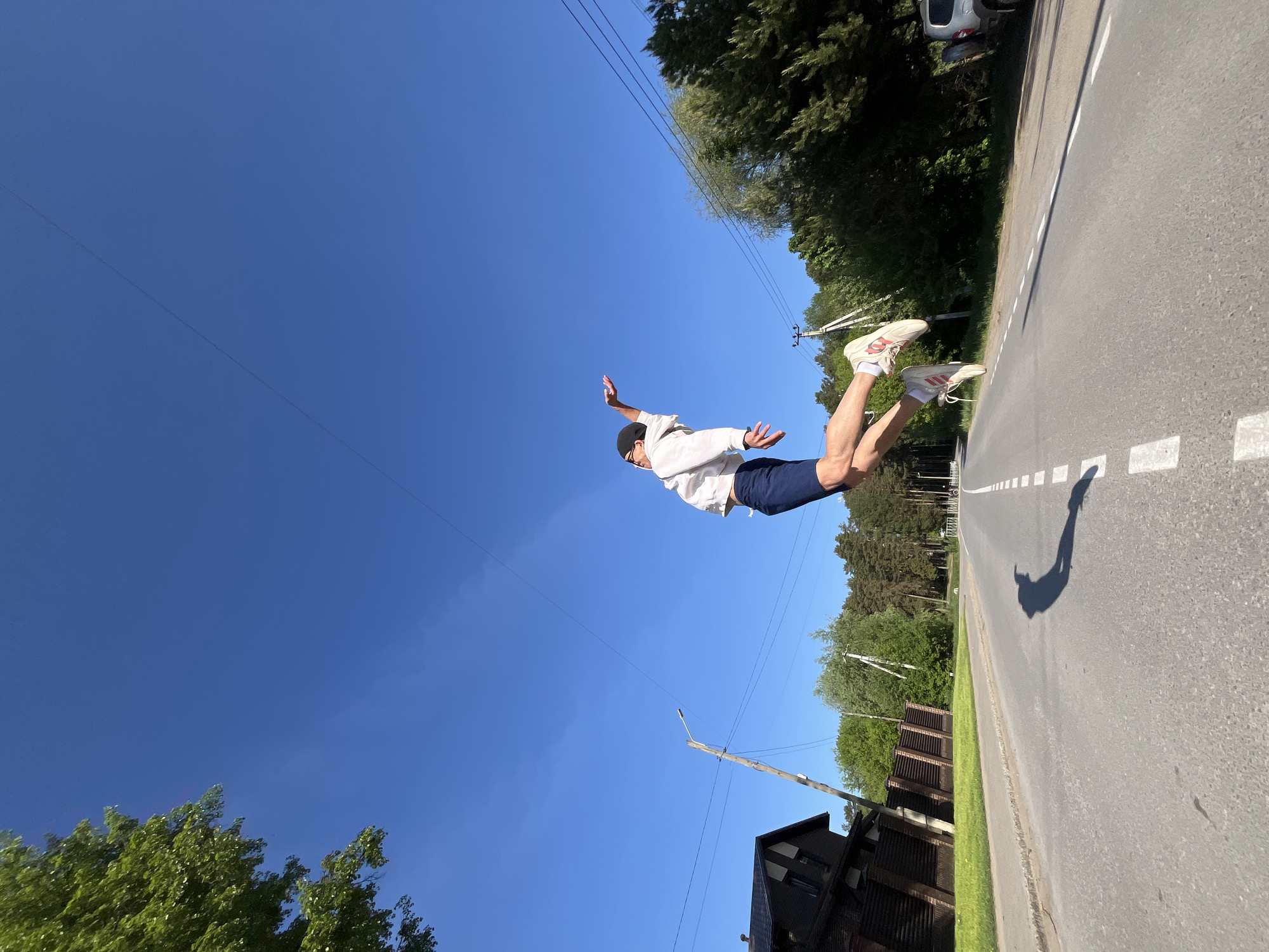A man appears to be floating horizontally above an asphalt road, casting a shadow on the road surface. He is wearing a white sweatshirt, navy shorts, and white athletic shoes with red accents. He has a dark cap on his head. The road has white painted lane markers. Trees and vegetation line one side of the road, along with a partial view of a dark-colored building. On the opposite side of the road, more trees are visible and a silver car is parked. Power lines stretch across a clear blue sky in the background. The location is Chaikovsky, Russia.