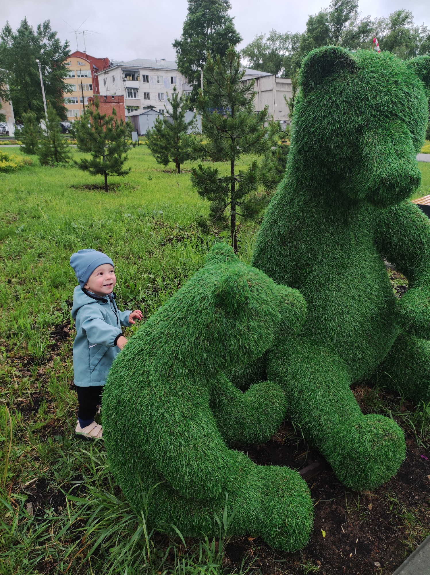 A child in a light blue jacket and matching beanie looks up at two large, green, grass-covered bear sculptures in a park. The child is standing on a patch of grass, and behind them, there are more young evergreen trees, grass, and buildings in the background. The location is the city of Chaikovsky, Russia.
