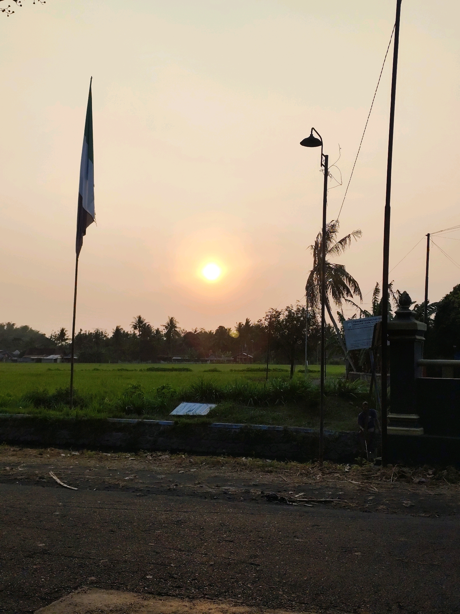 A wide-angle, slightly low-angle shot captures a sunset over a rural landscape in Bantul, Indonesia. The sky is a pale orange, transitioning to yellow near the horizon where the sun is visible as a bright, glowing orb. In the foreground, a dark, asphalt road shows signs of wear and scattered debris. To the left, a tall flagpole stands with a vertically striped flag, appearing to be green, white, and possibly black. Behind the flag, a field of vibrant green grass stretches towards a line of trees and scattered buildings. A prominent palm tree, silhouetted against the sky, stands near a dark, ornate gate. A street lamp with a curved arm is positioned on a pole to the right of the flag. A person, wearing a dark shirt and pants, is visible near the gate, facing away from the camera, and holding what appears to be a long stick. The overall scene evokes a peaceful, late afternoon atmosphere.