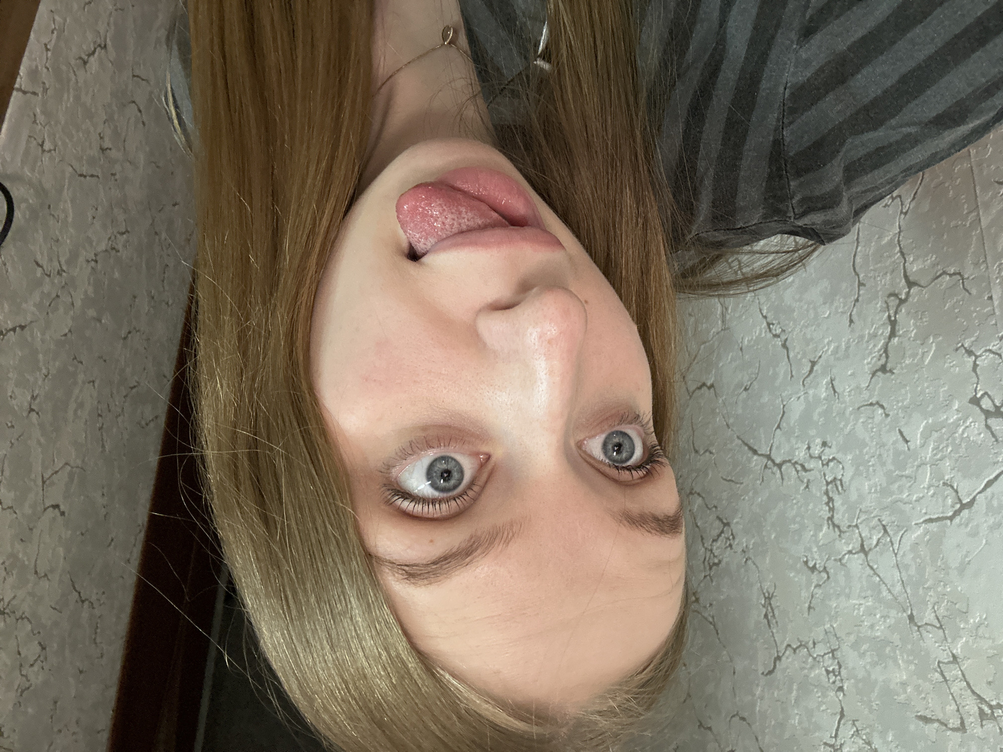A woman with light skin, blonde hair, and blue eyes is sticking out her tongue in the image. She is wearing a gray and black striped shirt. The background appears to be a light-colored wall with a cracked or marbled pattern. There's a door frame in the bottom left corner. The image is rotated so the woman appears upside down. The location of the city of Chaikovsky, Russia is not determinable from the image.