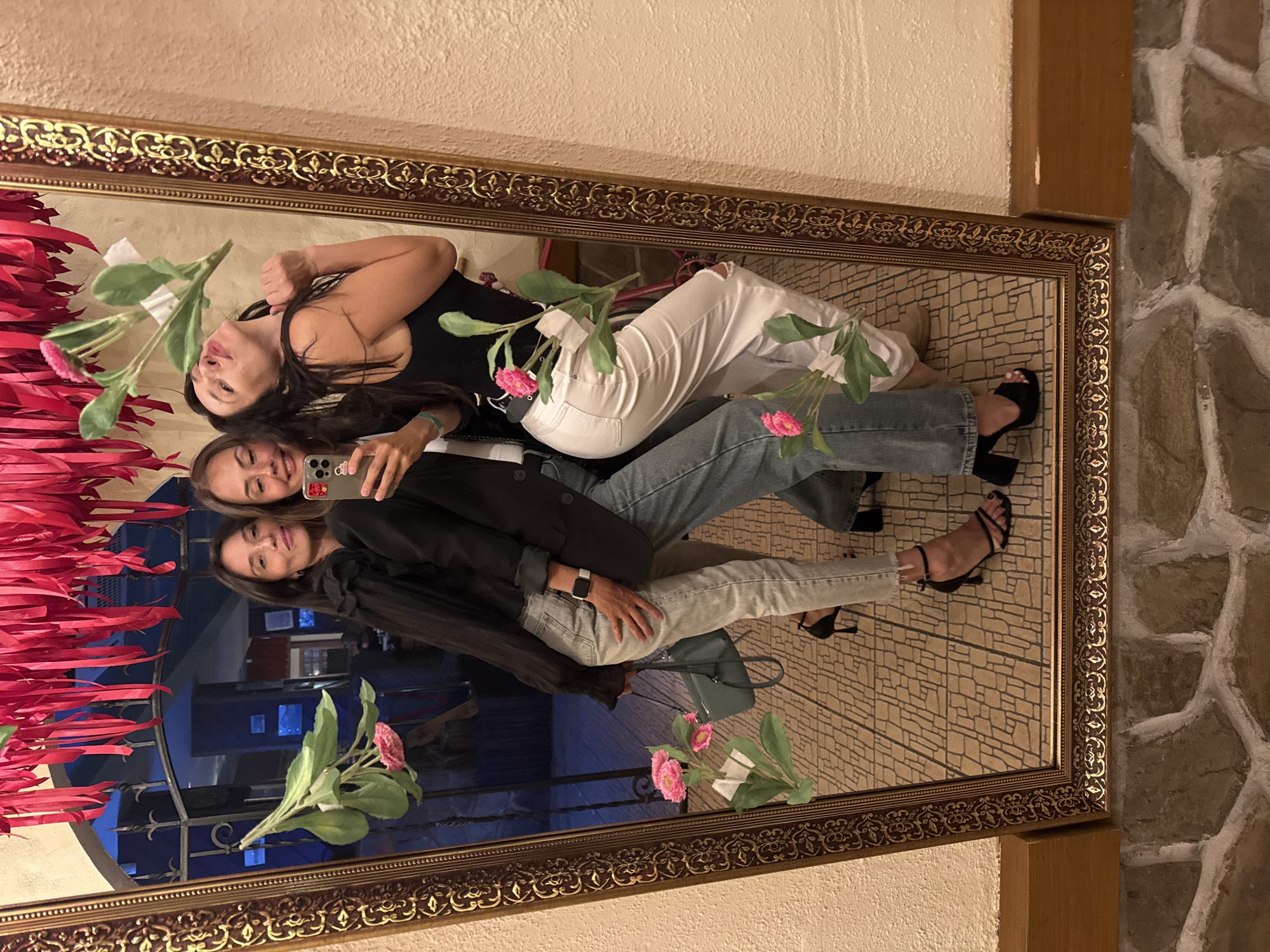 Two women are posing for a picture in a large, ornate mirror, seemingly indoors. The mirror reflects a scene with pink paper decorations and an exterior with a blue glass facade. The women are dressed in contemporary casual wear. One woman is wearing white jeans and a black top, while the other is wearing light-wash denim jeans and a dark blazer. They are holding a smartphone. The mirror's reflection suggests a public space, possibly a restaurant or venue with decorative elements. The location appears to be Chaikovsky, Russia, based on the background reflected in the mirror.