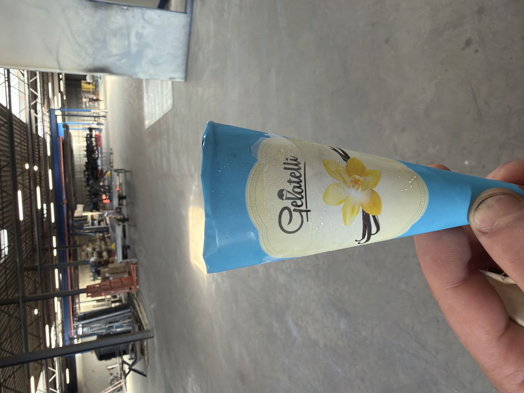 The image shows a hand holding a vanilla ice cream cone. The cone is wrapped in blue and yellow packaging with the brand name "Gellati" clearly visible in a stylized font. A yellow vanilla flower and two vanilla pods are depicted on the packaging. The cone appears to be slightly melting, with small droplets of condensation on the wrapper.
The background is a blurry industrial setting, likely a warehouse or factory, with metal shelving, lighting fixtures, and various objects stored on the shelves. The floor is a concrete surface. The lighting suggests it is daytime. The image appears to be taken in Mol, Belgium, as indicated by the location context. The hand holding the cone is the only visible person in the image. The overall impression is casual and informal, possibly indicating a break or a moment of personal enjoyment.