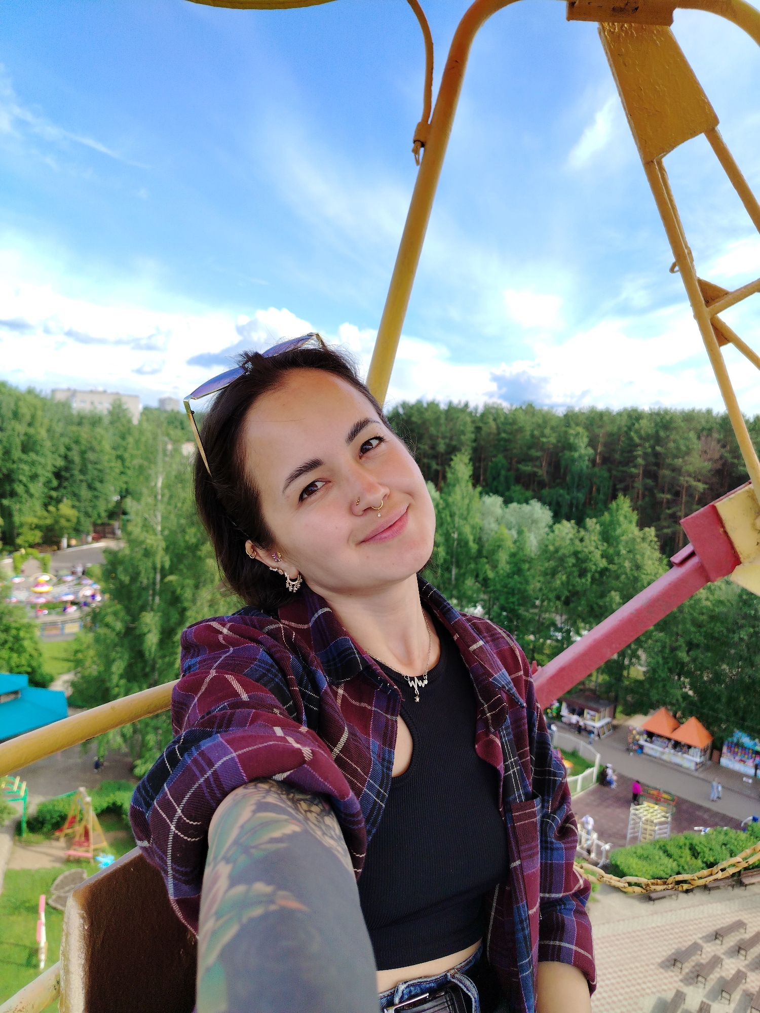 A young woman is depicted in a selfie, taken from a high vantage point, likely on a Ferris wheel, overlooking an amusement park and surrounding landscape. She has dark hair, brown eyes, and a pleasant, gentle smile directed at the camera. Sunglasses rest on top of her head. She wears a dark red, blue, and purple plaid button-up shirt over a black ribbed crop top. Her visible left arm features a colorful sleeve tattoo. Notable facial piercings include a nose ring (hoop with a small gem), a septum piercing, and multiple earrings. A silver-colored necklace with a cursive "MIMI" pendant is visible around her neck.
The background reveals a sunny daytime scene with a bright blue sky and scattered white clouds. Below, the yellow and red metal structure of the Ferris wheel frames the view. The park features green grassy areas, paths, and distant amusement rides such as a carousel and swings. Beyond the park, a dense forest stretches out, and in the far distance, several multi-story residential buildings are visible. The overall setting suggests a vibrant summer day at an amusement park in Chaikovsky, Russia.