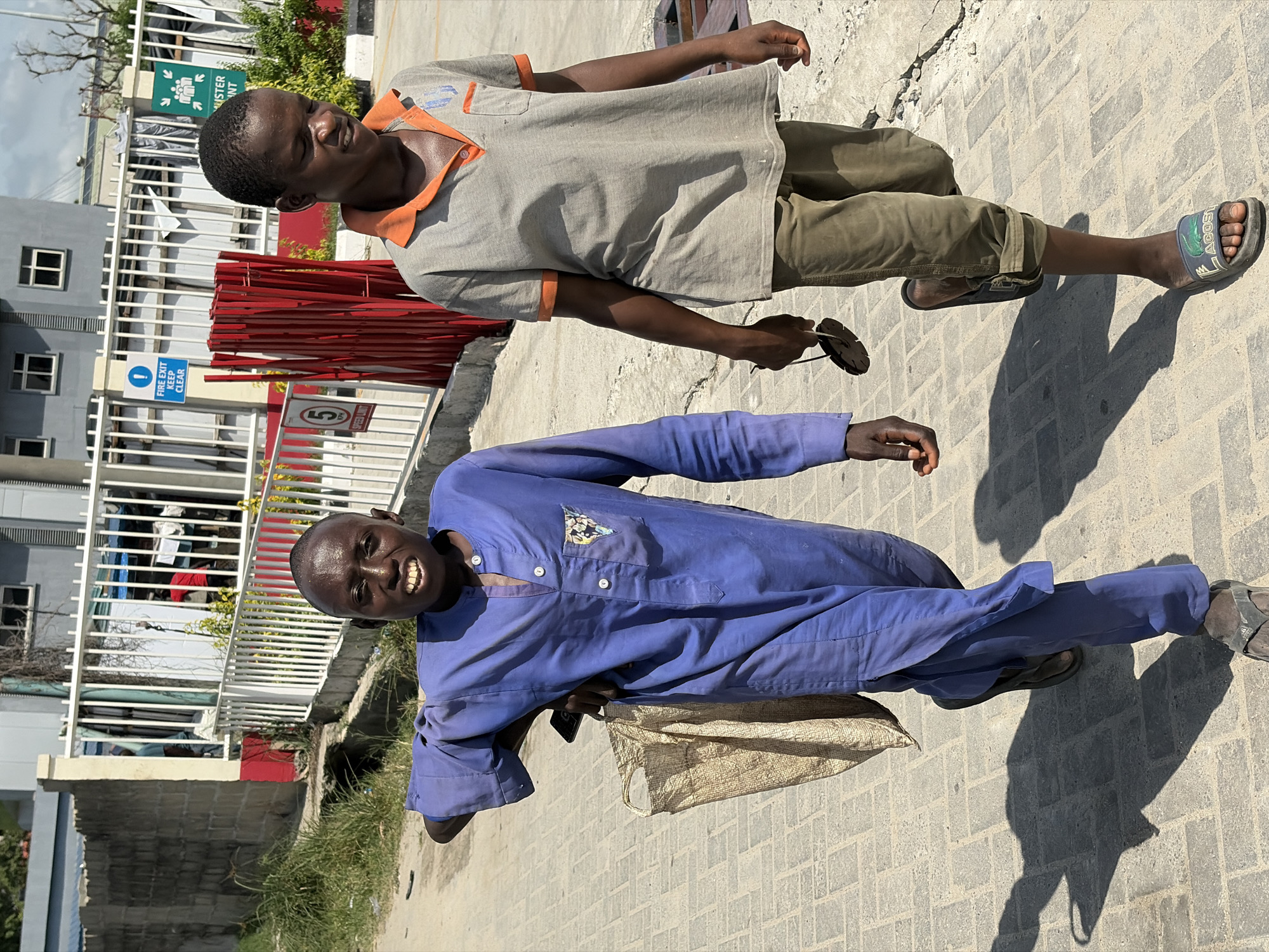 Two men are captured in a bright, sunlit outdoor scene on a paved walkway in Igboefon, Nigeria. The strong sunlight creates distinct shadows, indicating a clear, sunny day.
In the foreground, a man wearing a long-sleeved blue uniform-like outfit with a patterned pocket smiles broadly at the camera. He carries a light-colored woven bag and holds a mobile phone in his left hand, wearing dark sandals.
Slightly behind him and to the right, another man wears a grey short-sleeved polo shirt with orange trim, olive green cargo pants, and green and black Lacoste-branded slides. He holds a pair of sunglasses in his right hand and looks towards the left with a neutral expression, possibly squinting in the bright sun.
The ground consists of grey interlocking paving bricks. The background features various urban structures: a building with white railings, a multi-story grey building with windows, and a stack of red metal bars or stakes. Green foliage and a grassy area are also visible.
Several signs are present: a blue sign with an exclamation mark stating "FIRE EXIT KEEP CLEAR," a white sign displaying "(5) SPEED LIMIT," and a partial green sign with text reading "STER INT" and "M". The scene depicts an everyday moment, possibly during a walk along a street or pathway.