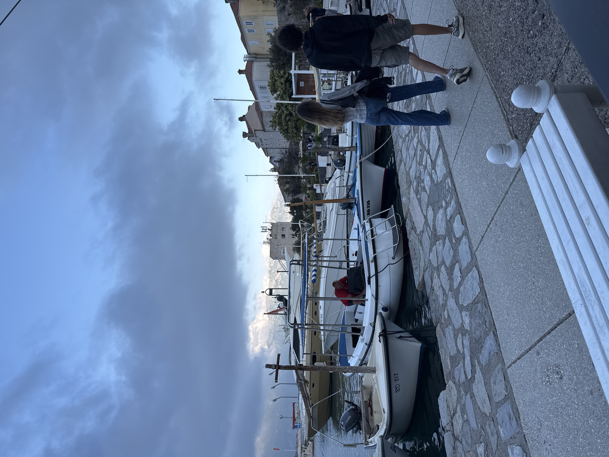 Two people are walking on a stone pathway alongside docked boats on a cloudy day in Budva, Montenegro. One person is wearing a black jacket and khaki shorts, while the other wears a plaid shirt and jeans. The pathway is lined with boats to the left, and a white bench with decorative posts to the right. In the background are buildings, trees, and mountains. The sky is overcast with dark clouds.